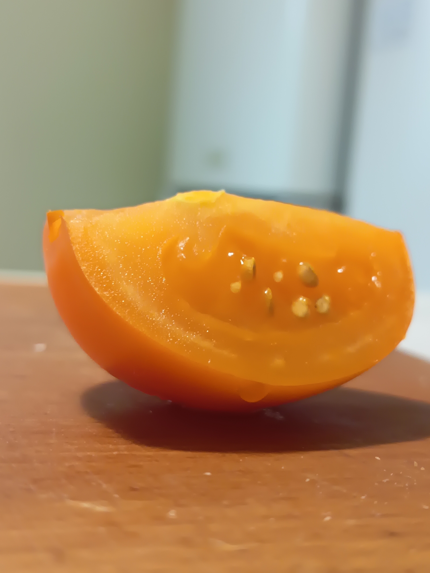 A close-up, eye-level shot focuses on a single, bright orange, halved cherry tomato resting on a wooden surface. The tomato is cut vertically, revealing its juicy interior with small, light-colored seeds suspended within the gelatinous pulp. A tiny droplet of juice is visible on the bottom edge of the tomato. The background is softly blurred, showing a light green wall on the left and a lighter, almost white vertical element on the right. The lighting creates a subtle shadow behind the tomato. The scene is indoors, likely in a kitchen, with no specific landmarks indicating the city of Izmail, Ukraine, or any discernible environmental features associated with that location.