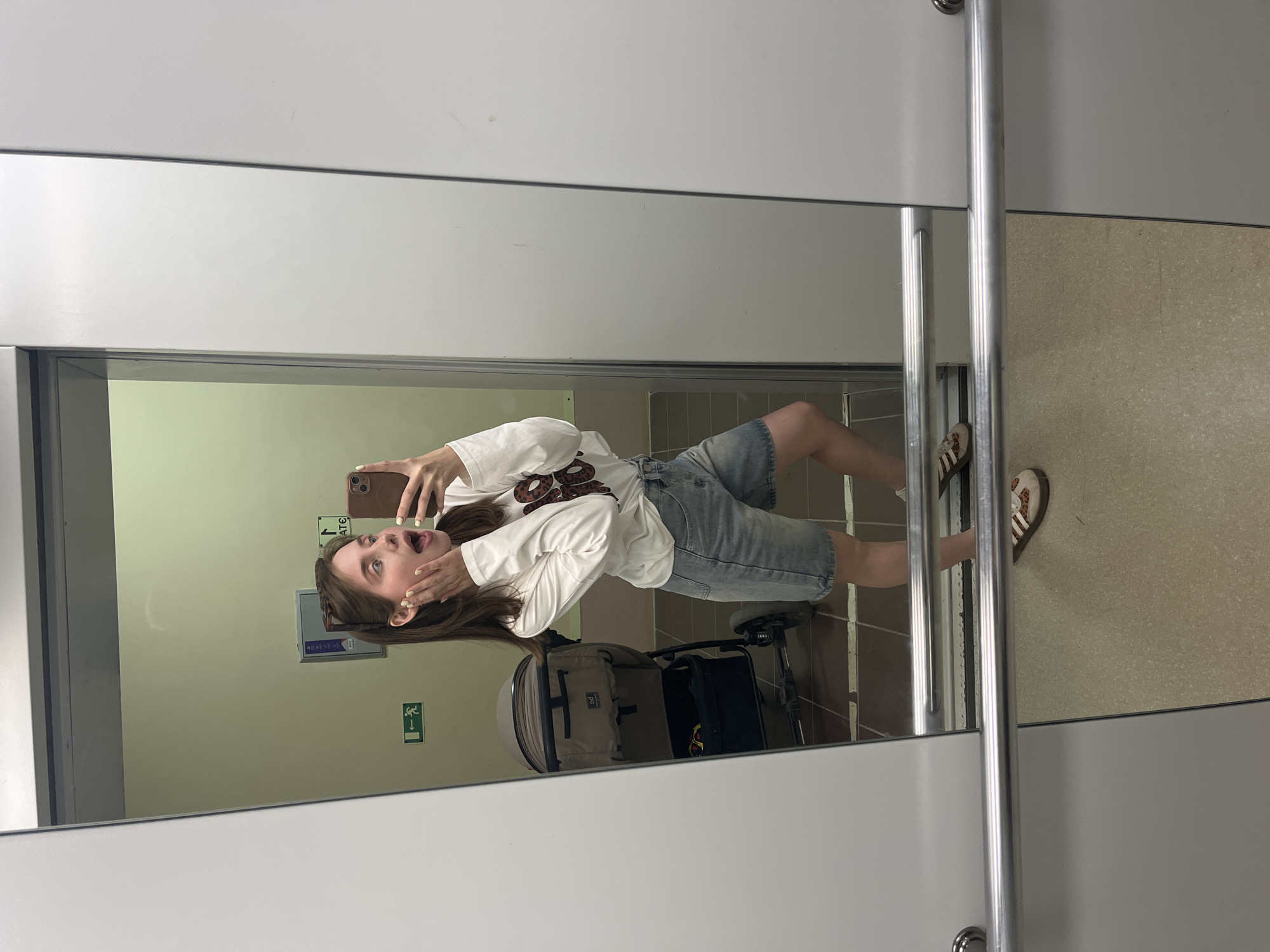 A young woman stands in a public restroom, possibly in Chaikovsky, Russia, based on environmental context. She holds a phone to take a selfie, expressing surprise or excitement with her hands framing her face and an open-mouthed expression. She is wearing a long-sleeved white shirt with a graphic design, denim shorts, and slides.
The restroom stall has light green walls with signage, including a green exit sign. The floor has brown tiling. A stroller is visible behind her, suggesting the restroom is in a family-friendly location. The stall door is white with metal hardware and framing. The overall lighting is bright and even, indicating modern restroom facilities.
