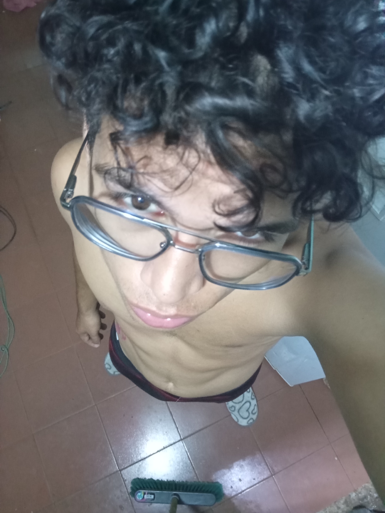 A person with dark, curly hair and wearing glasses is positioned in the center of the frame, looking up at the camera. The person is shirtless, revealing their torso. They are wearing dark-colored bottoms. The person is standing on a tiled floor. To the left of the person is a green cord. In the bottom of the frame, there is a green push broom. Behind the person is a white object. One slipper is visible. There is no indication that the photo was taken in the city of Altagracia, Venezuela.