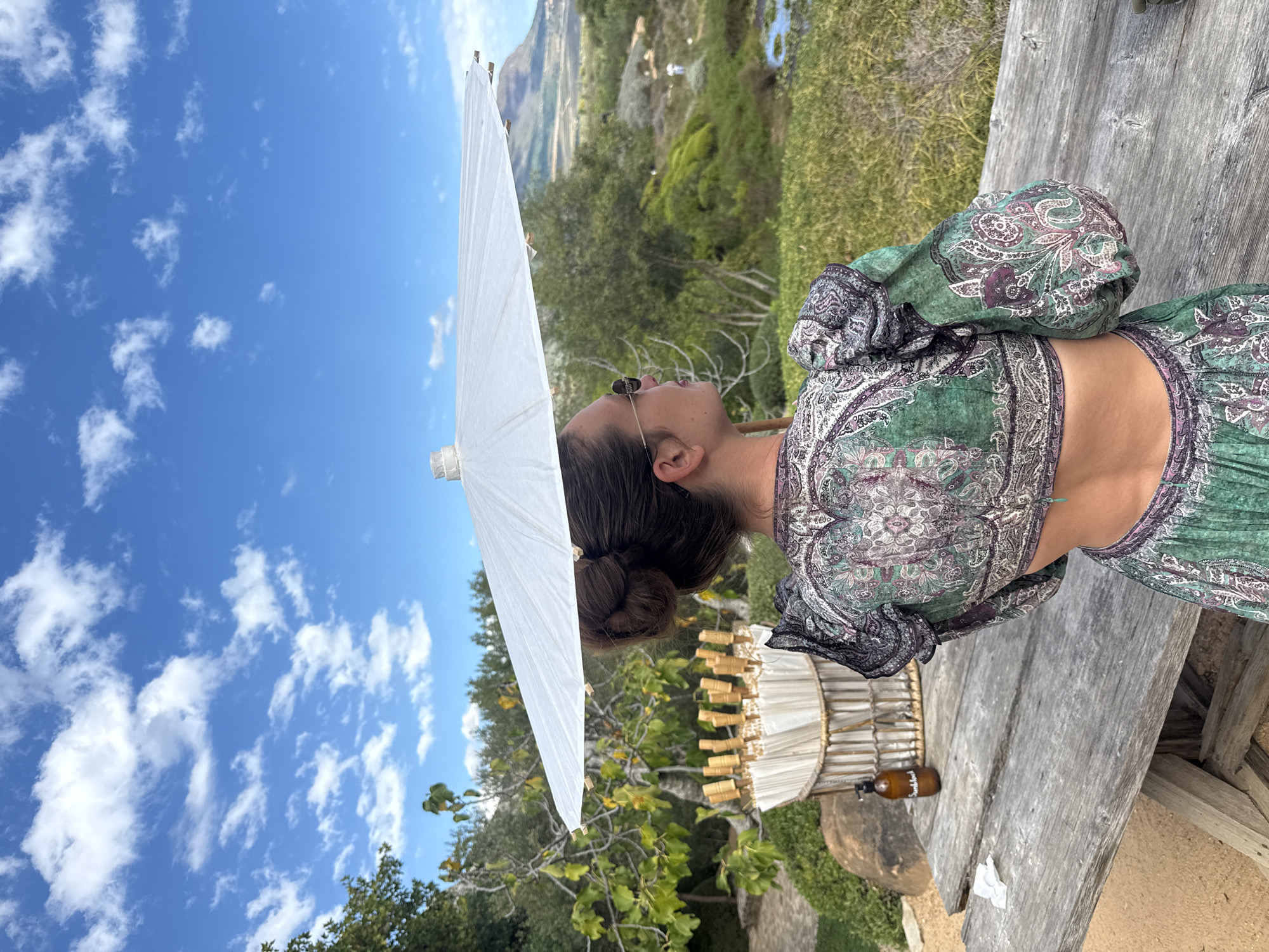 A woman with her hair in a bun wears sunglasses and a patterned two-piece outfit with long, loose sleeves. She is holding a white parasol in front of her, the sky is blue with scattered clouds and there are trees and hills in the background. The woman is sitting at a weathered wooden table next to what appears to be a lantern and small brown bottle. The scene appears to be taken outdoors with natural lighting in Stellenbosch Nu, South Africa.