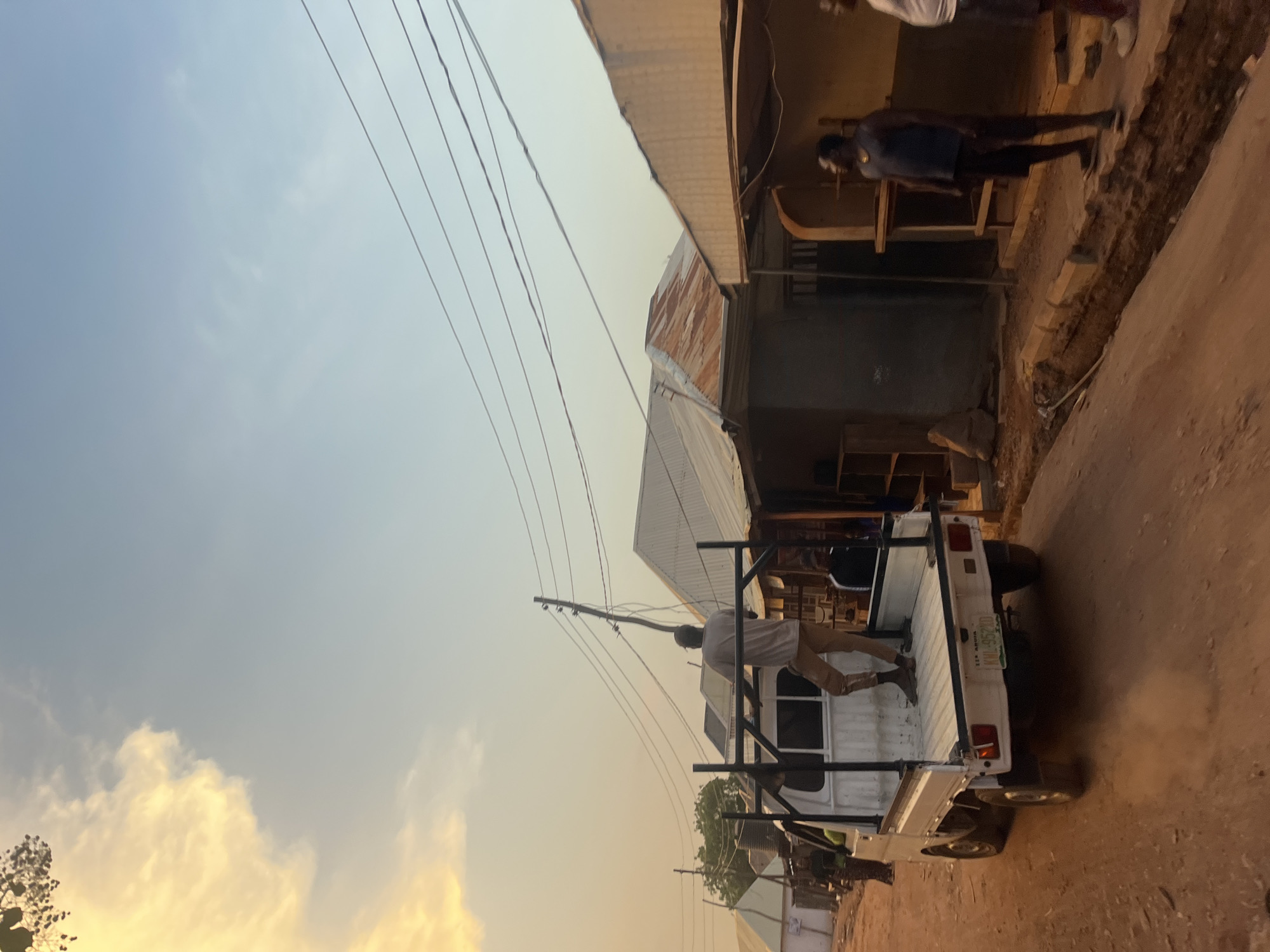 The image, taken at dusk in Abuja, Nigeria, shows a white pickup truck on a red dirt road alongside weathered buildings. A man in a light-colored shirt and dark pants is on the back of the truck, near the metal structure that's affixed. Electrical wires are visible overhead against the sky. Another man is standing on the ground next to an outdoor space, and there appears to be carpentry work happening there. The sky is a gradient of blue and orange. The license plate reads "KUL 552ZD" above the text "SEE NIGERIA."