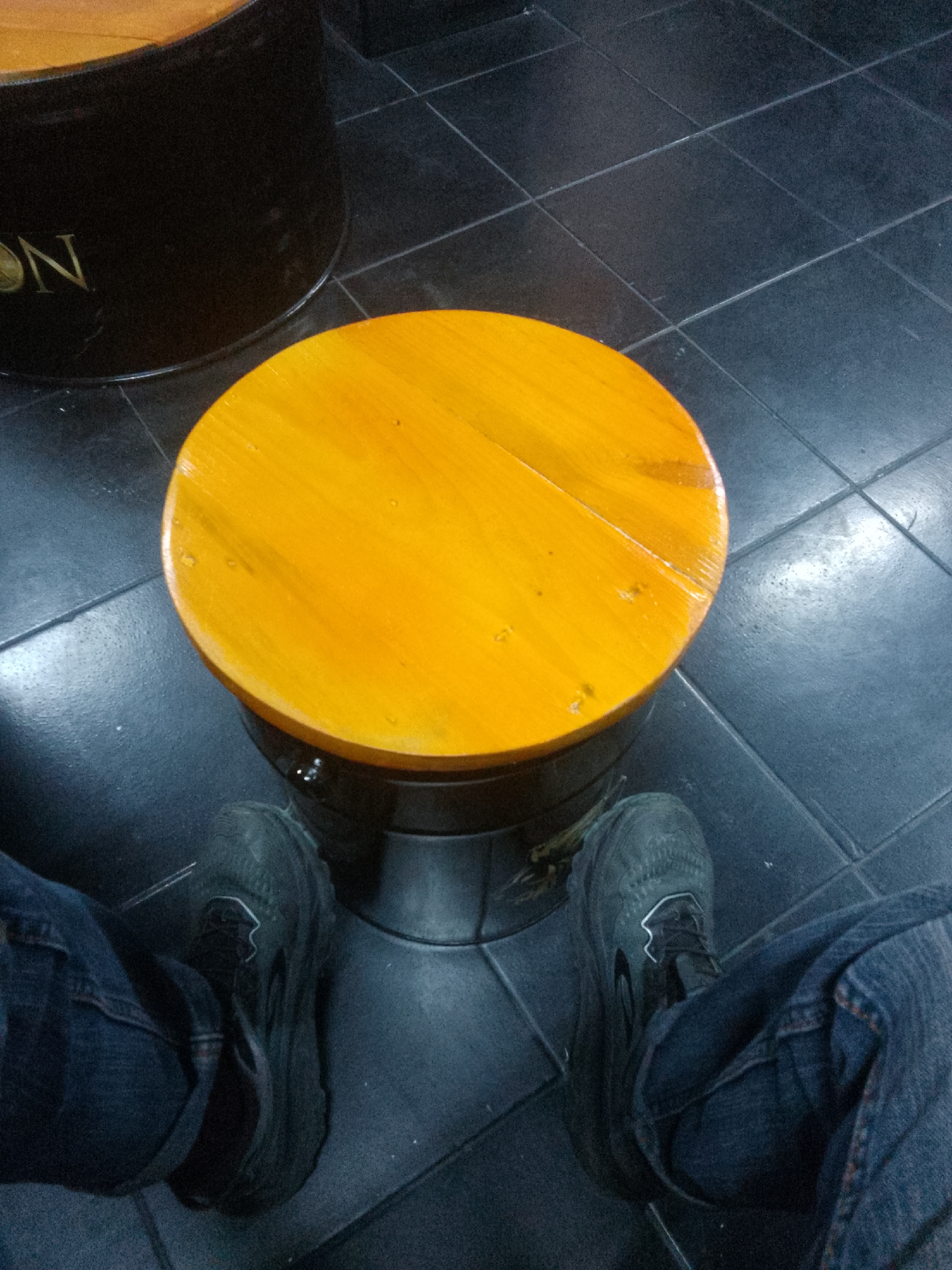 The image shows a high-angle shot of a person's legs and feet, wearing dark blue jeans and dark athletic shoes, sitting on a wooden stool. The stool has a round, orange wooden top and a dark, cylindrical base. In the background, a portion of a dark, cylindrical object with gold lettering is visible, along with a dark tiled floor. The scene appears to be indoors. The lighting is dim, with reflections visible on the stool and floor. No specific landmarks or context are provided to indicate a location in Trenggalek, Indonesia.