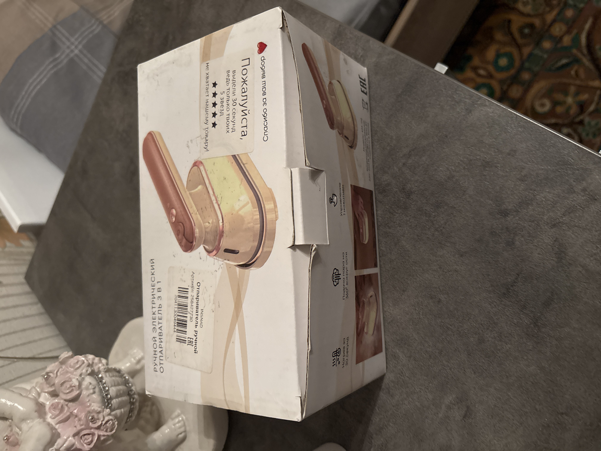 This image captures a white cardboard box containing a handheld electric steamer, described as a "3-in-1" device, situated on a dark gray, textured surface in Chernogorsk, Russia. The box features an image of the rose-gold/copper-colored steamer and Russian text. Visible text includes "РУЧНОЙ ЭЛЕКТРИЧЕСКИЙ ОТПАРИВАТЕЛЬ 3 В 1" (Handheld Electric Steamer 3 in 1), an article number "258427730," and a barcode. Other texts, some oriented upside down, request a "5 STARS" review, stating "Пожалуйста, Спасибо за ваш выбор" (Please, Thank you for your choice) with a red heart. Product features mentioned are "Нагрев за 30 секунд" (Heating in 30 seconds), "Подача пара на 360° вокруг оси" (Steam delivery 360° around the axis), and "Управление 1 нажатием" (One-button control). An "EAC" conformity mark is also present. To the left, a white ceramic figurine, possibly an angel or cherub holding a basket of pink roses with silver embellishments, is partially visible. The background is blurred, showing light-colored furniture or a wall and a patterned textile, possibly a cushion or blanket, with gray and brown stripes. A small portion of a patterned rug or carpet in shades of brown, green, and gold is visible on the far right.