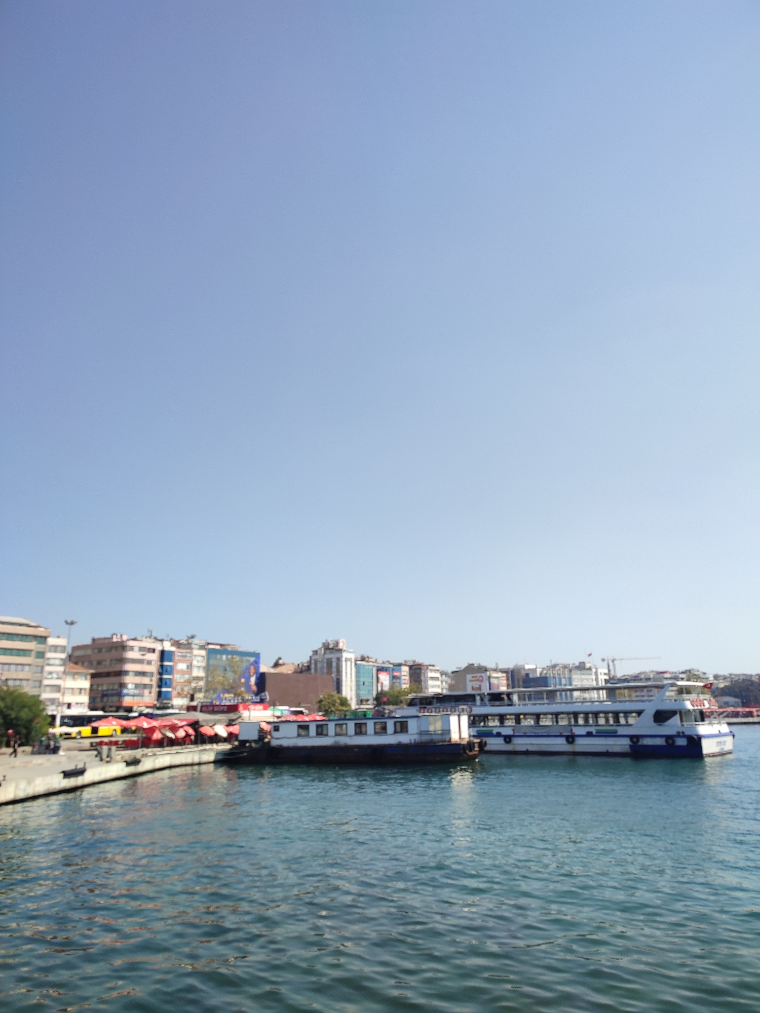 The image shows a waterfront scene in Kadıköy, Turkey, with several ferries docked along the shore. The sky is a clear blue, and the water is calm, with gentle ripples. In the background, a line of buildings with various architectural styles forms the Kadıköy skyline. Some of the buildings appear to be residential, while others seem to be commercial establishments. Red umbrellas, possibly from market stalls or cafes, are visible along the waterfront. The ferries are large passenger vessels, with multiple decks and windows. One ferry in the foreground is white with a blue stripe along its hull, and it appears to be docked next to another, darker vessel. The overall impression is of a bustling urban waterfront on a sunny day.
