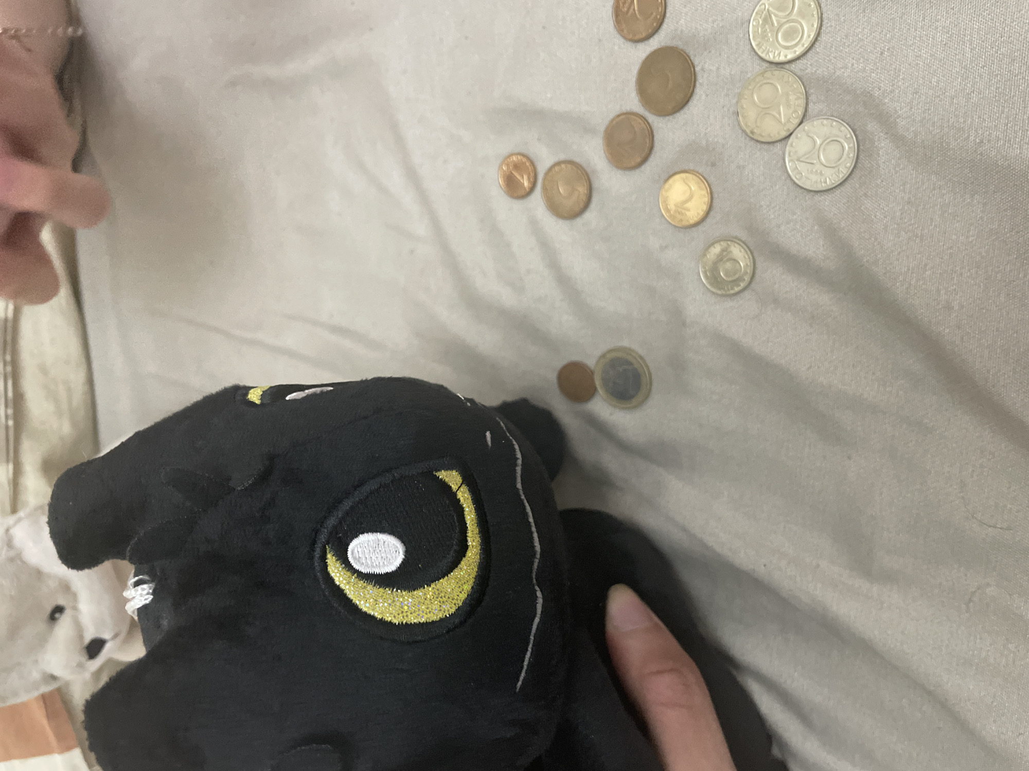 An indoor scene, presumably a residential setting in the city of Ihtiman, Bulgaria, displays a collection of objects on a light grey, textured fabric surface, likely bedding. In the foreground, a black plush toy resembling Toothless from "How to Train Your Dragon" is positioned, with a human hand holding its lower left side. The plush features prominent yellow glitter eyes with white pupils and a stitched white line indicating a mouth. In the upper background, partially obscured and out of focus, a white plush toy, possibly a bear, is visible. Adjacent to the white plush, a child's hand with a thin gold-colored chain bracelet is present, reaching into the frame from the upper right. Scattered across the light grey fabric surface are various coins. These include one 1 Euro coin, one gold-colored 1 Bulgarian Lev coin, several copper-colored coins (likely Euro cents or Bulgarian stotinki), and several silver-colored Bulgarian stotinki coins, including denominations of 20 and 50, with discernible Cyrillic text. The image provides no visual cues to directly depict the city of Ihtiman, but the presence of Bulgarian currency is consistent with the specified Bulgarian location.