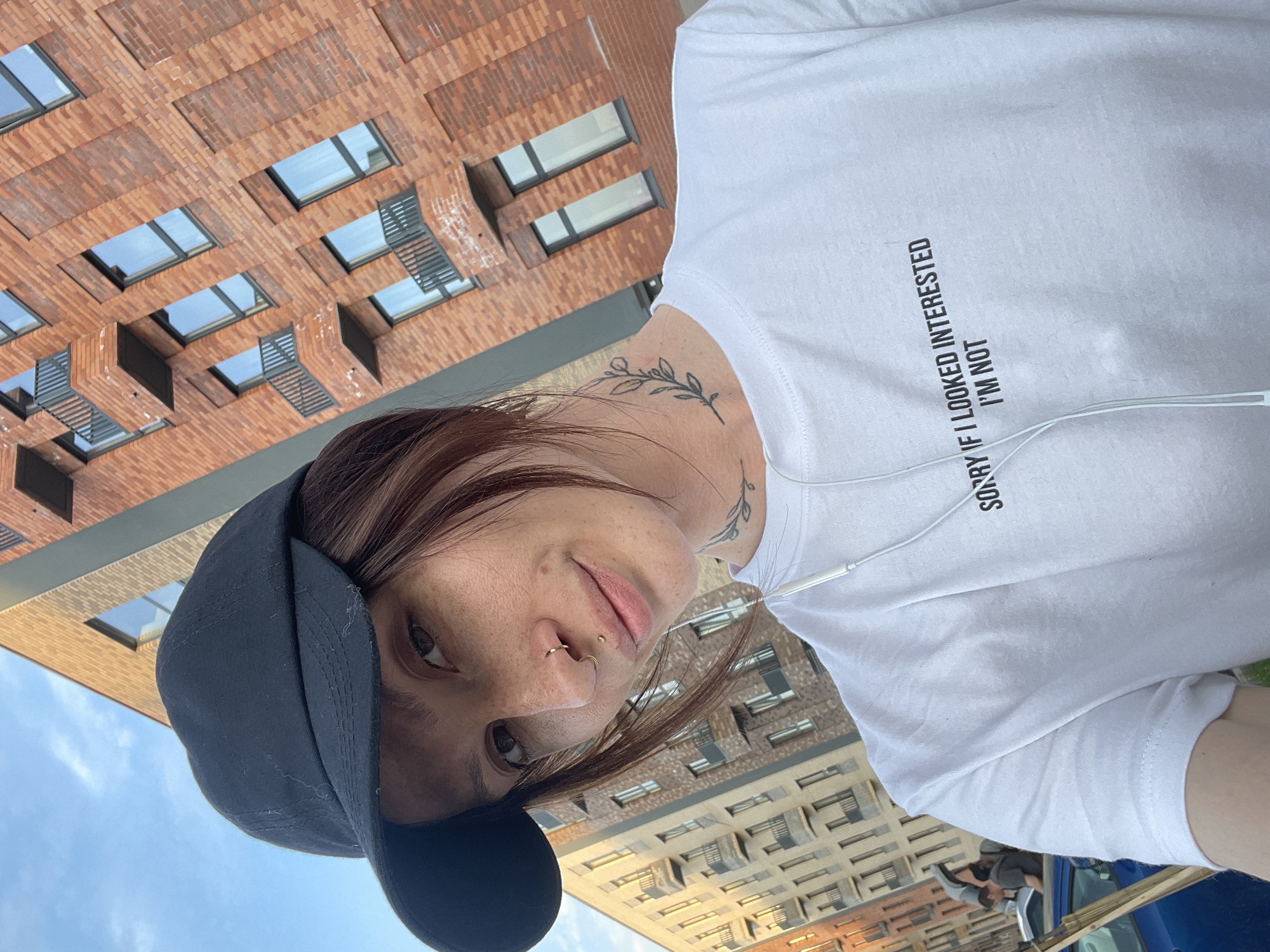 A person wearing a dark cap and a white t-shirt with the phrase "SORRY IF I LOOKED INTERESTED I'M NOT" is in an outdoor environment. The person has a nose ring and a neck tattoo. In the background, there are brick buildings with windows, possibly in a city environment, potentially Izhevsk, Russia. Other people and a blue car can be seen further in the background.