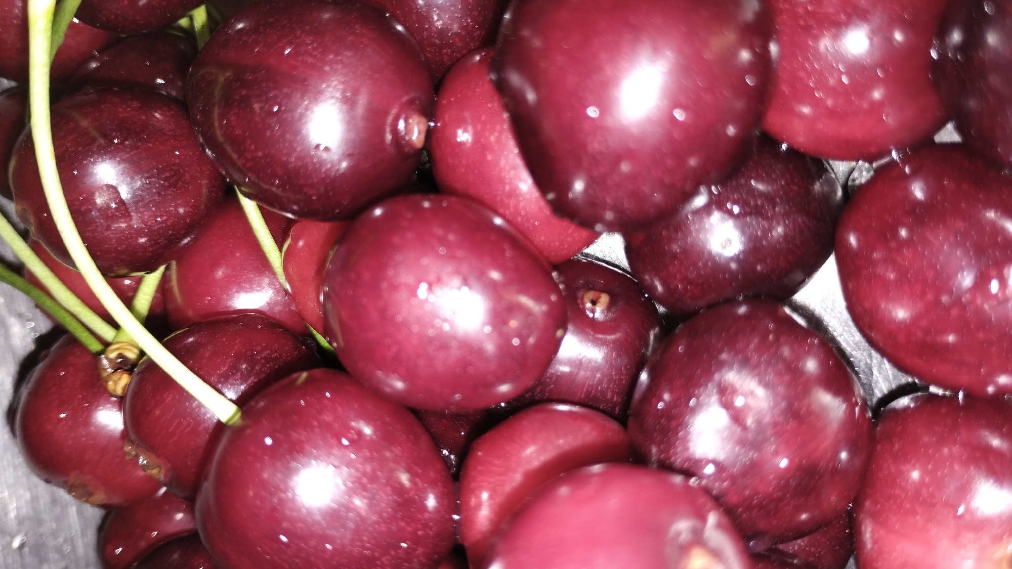 A close-up, overhead shot shows a pile of fresh, dark red cherries. The cherries are plump and glistening, with small water droplets visible on their surfaces. Several stems are intertwined amongst the fruit, some with a lighter green hue. The lighting casts bright highlights on the cherries, suggesting a reflective surface. The image is a macro photograph, focusing on the texture and color of the fruit. The background is dark and out of focus, drawing all attention to the cherries. There are no people or environmental elements present beyond the fruit itself.