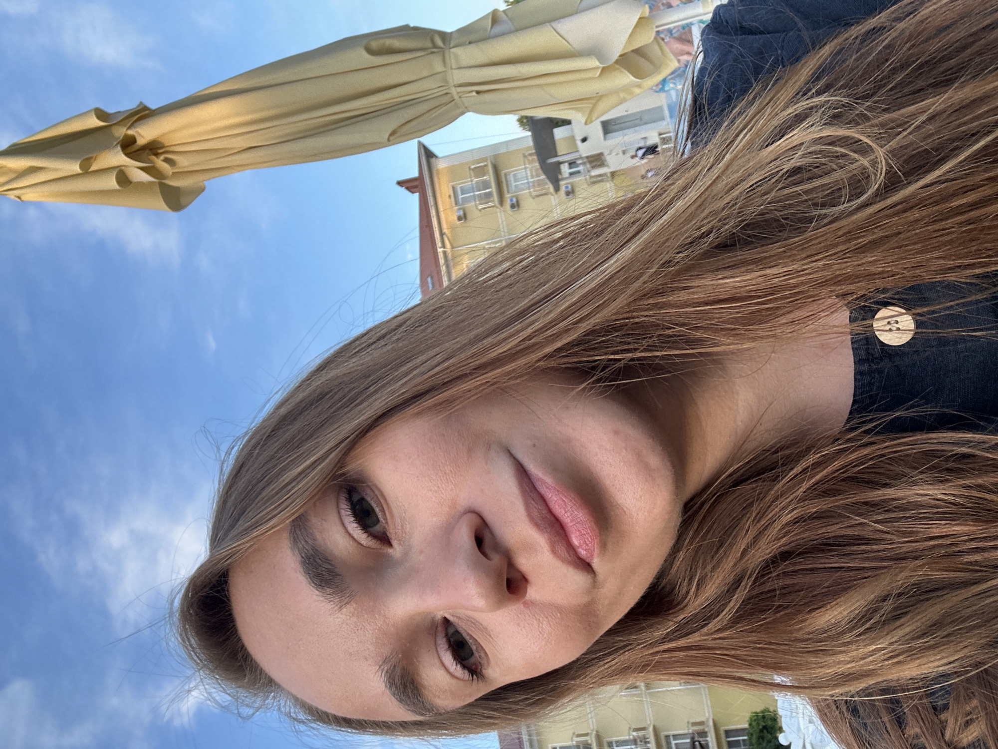 A young woman with long, light brown hair takes a selfie outdoors with a blue sky and some clouds above. She is wearing a dark shirt with a wooden button on the front. In the background, a yellow building with multiple balconies and air conditioning units is visible. To the right of the woman, a closed yellow umbrella is leaning upright, and part of a beach or resort area with lounge chairs and people can be seen in the distance. The scene appears to be set in Vityazevo, Russia, a coastal resort town on the Black Sea.