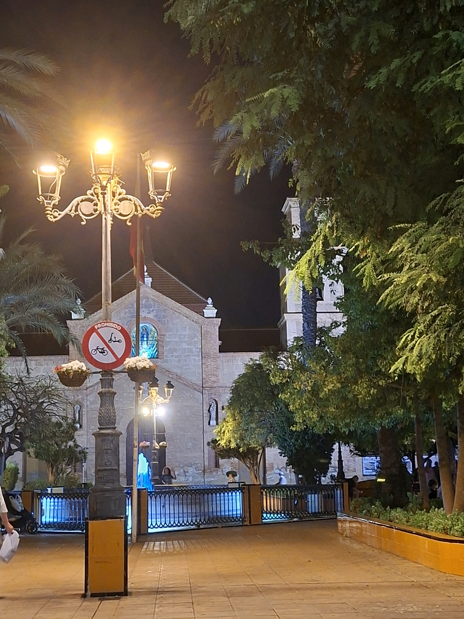 The image captures a nighttime scene in Torrevieja, Spain, likely in a town square or pedestrian area. A multi-lamp lamppost, ornately decorated and emitting a warm yellow light, stands prominently in the foreground. Attached to the lamppost is a circular sign with a red border, indicating a prohibition. Inside the circle, silhouettes of a scooter and a bicycle are crossed out. Two hanging flower baskets are affixed to the lamppost, containing white flowers.
Behind the lamppost, a building with a light-colored brick facade and a tiled roof is visible. It appears to be a church or similar public building, with a stained-glass window featuring a blue depiction of a figure. A bell tower with a distinct arched opening rises behind the main structure. Trees with dense green foliage are scattered throughout the scene, some illuminated by the artificial light.
In the mid-ground, a low black metal fence with decorative elements encloses an area with blue lighting. Beyond the fence, several people are present, some interacting near a fountain that emits a bright blue glow. The ground is paved with light-colored square tiles, reflecting the ambient light. The overall atmosphere is tranquil and illuminated, suggesting a public space that is active in the evening.