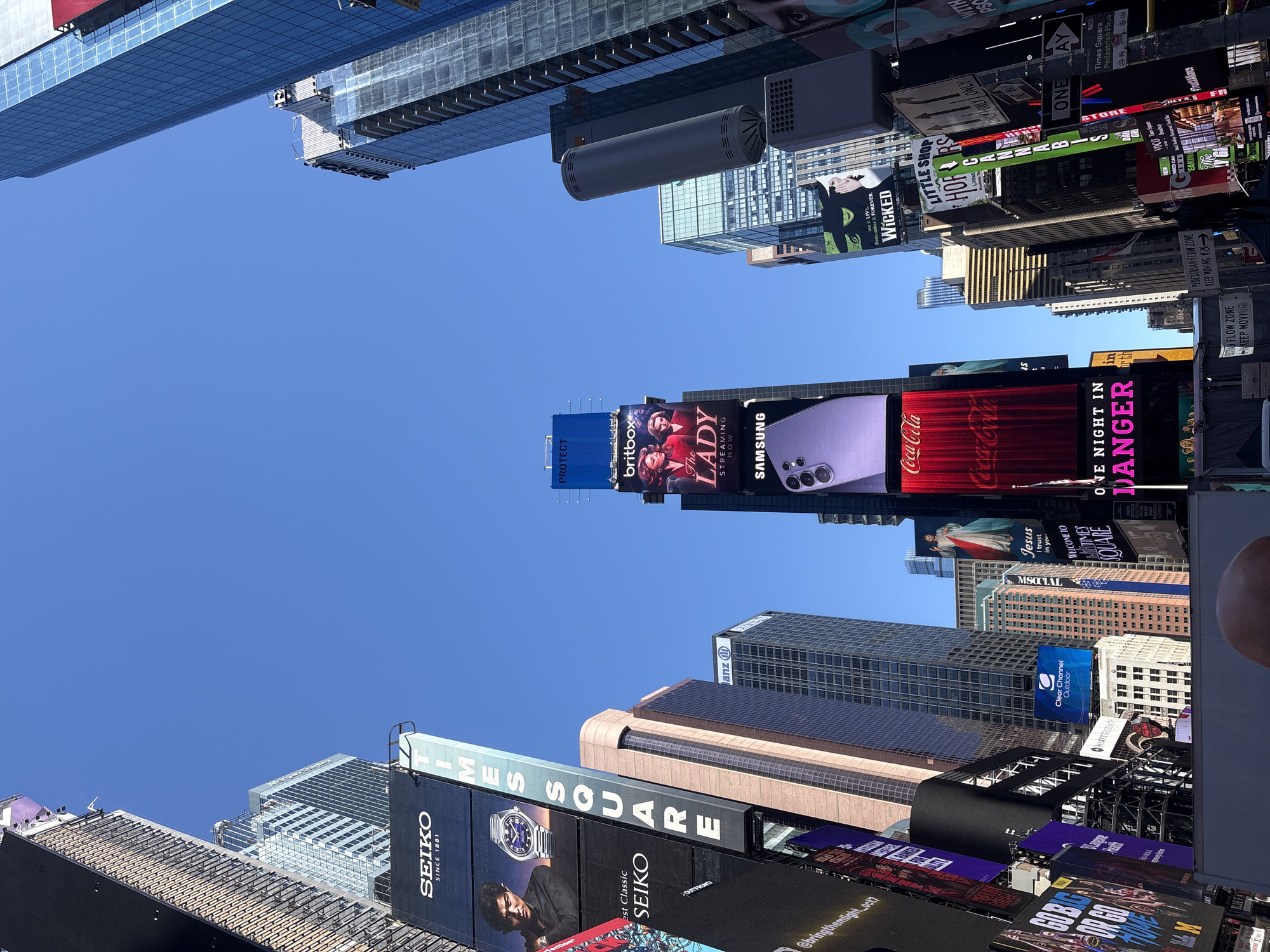 The image depicts a view of Times Square in New York City, United States of America. The perspective is angled upward, showcasing the towering buildings adorned with numerous digital billboards displaying advertisements for various brands and shows, including Seiko, Times Square, ANZ, Britbox's "The Lady," Samsung, Coca-Cola, Wicked, and others. The sky above is clear and blue. The overall scene captures the vibrant and busy atmosphere of Times Square.