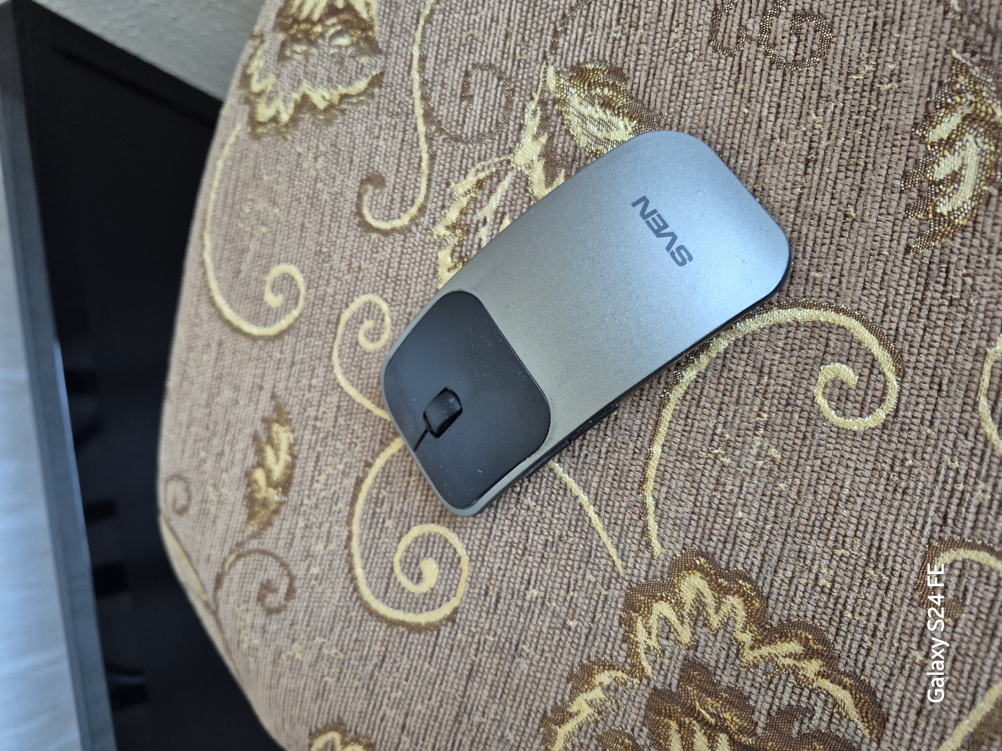 The image shows a silver and black computer mouse with the brand name "SVEN" visible on its surface. The mouse is placed on a textured, brown fabric surface with an ornate yellow pattern. The background is blurred, but appears to be a piece of furniture and a wall. The bottom left corner of the image contains a watermark that reads "Galaxy S24 FE". The image does not contain any information that would allow for determining its location in Chișinău, Moldova.