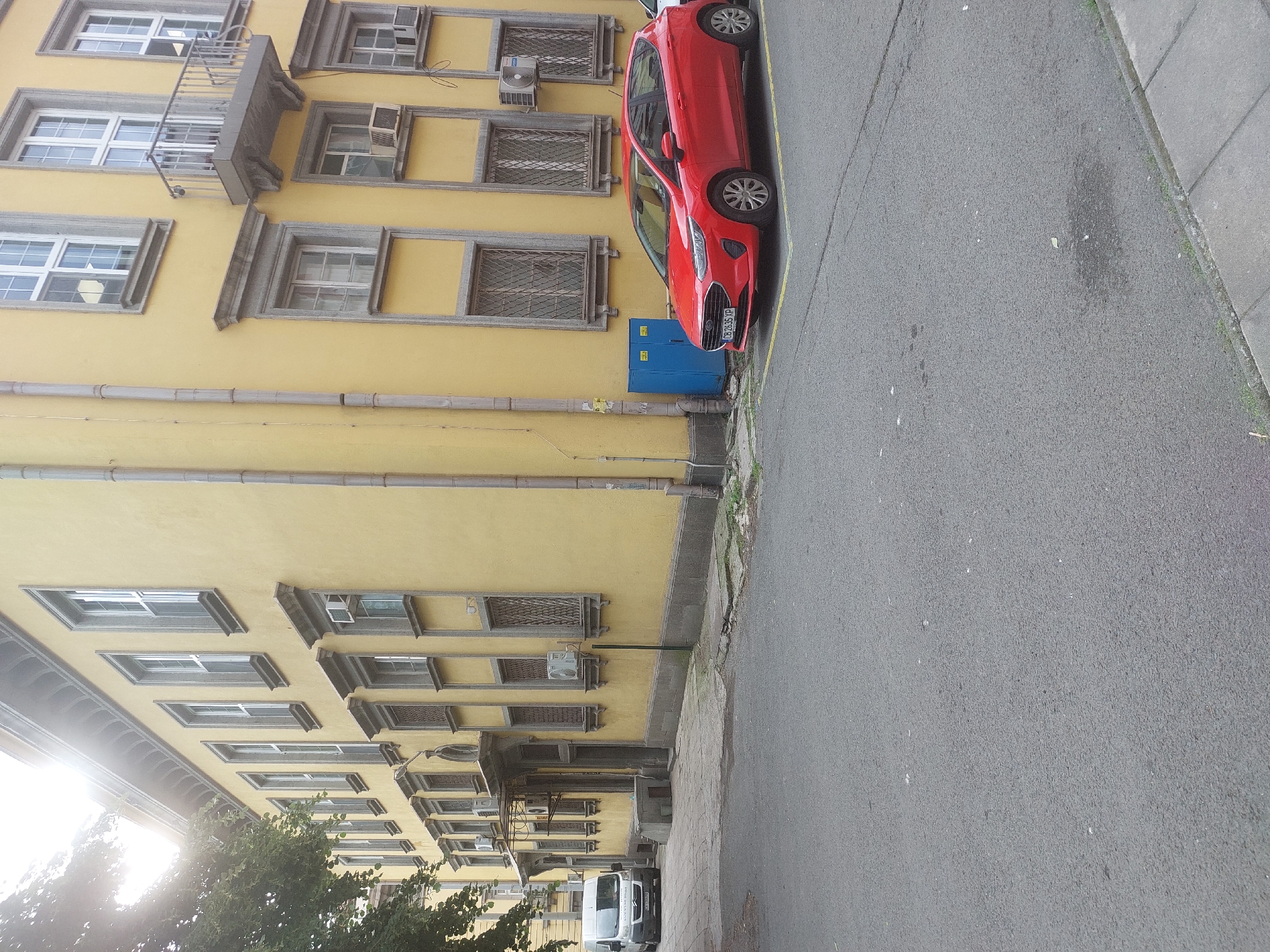 The image depicts a street scene in Burgas, Bulgaria. A red car is parked alongside a yellow building with ornate window frames, some with grills and air conditioning units. A blue electrical box sits near the car. The street is paved and a sidewalk runs along the right side of the frame. Further down the street, trees and a white van are visible. No people are visible in the frame. The license plate of the red car is visible.