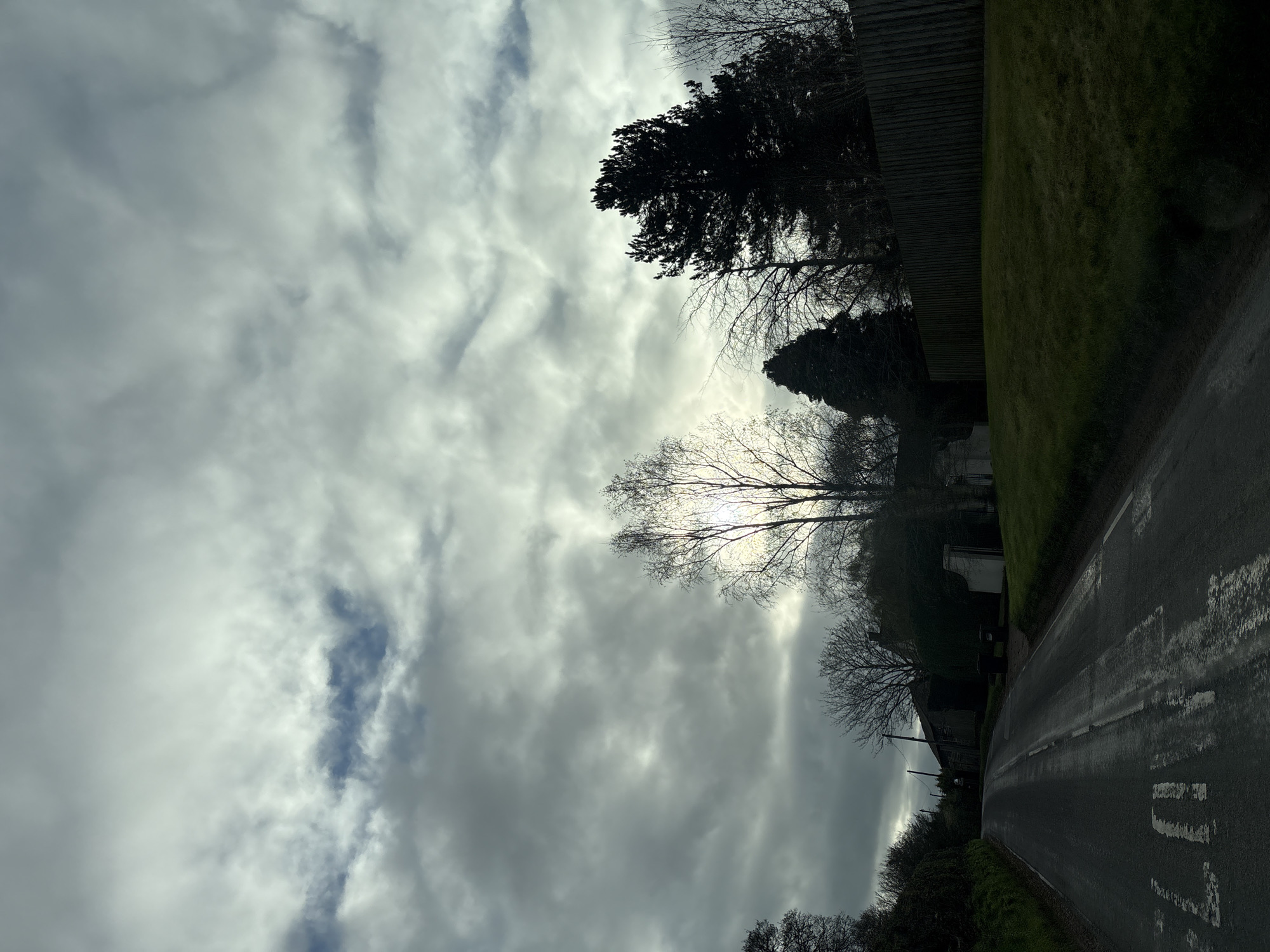 A sideways-oriented, overcast landscape view shows a wet road winding between a green grassy verge and a wooden fence on the right, with the sky dominated by a cloudy, bright sky. Leafless trees are silhouetted against the sky. On the left side of the road, there are a few buildings, including what appears to be a house or barn. The road has white markings. Given the available information, it is not possible to identify the exact location within Ruyton-XI-Towns, United Kingdom, without further details.