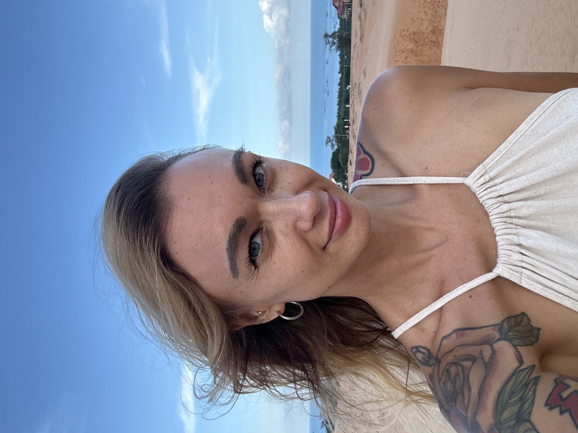 The image is a selfie of a young woman, rotated 90 degrees clockwise, depicting her with a bright, relaxed expression. She has blonde hair with darker roots, blue-grey eyes, and fair skin. She wears a light-colored, possibly off-white or cream, ruched-front top with thin spaghetti straps. A silver hoop earring and a smaller stud piercing are visible on her right ear.
Numerous tattoos are visible on her arms and shoulders. On her left upper arm/shoulder, a large floral tattoo, primarily roses with green leaves, covers a significant area. A distinct red and black cloud-like symbol, resembling the Akatsuki cloud, is visible on her right shoulder strap area.
The background shows a clear blue sky with sparse white clouds at what would be the top of a normally oriented image. To her right, a wide expanse of ocean or a large bay stretches towards the horizon, dotted with several boats. Beyond the water, a distant landmass with vegetation and some buildings is visible. Immediately behind her on the left side of the image, there's a sandy, earthy terrain, possibly a dune or hillside, consistent with a sunny coastal region like Mũi Né, Vietnam. There is no visible text.