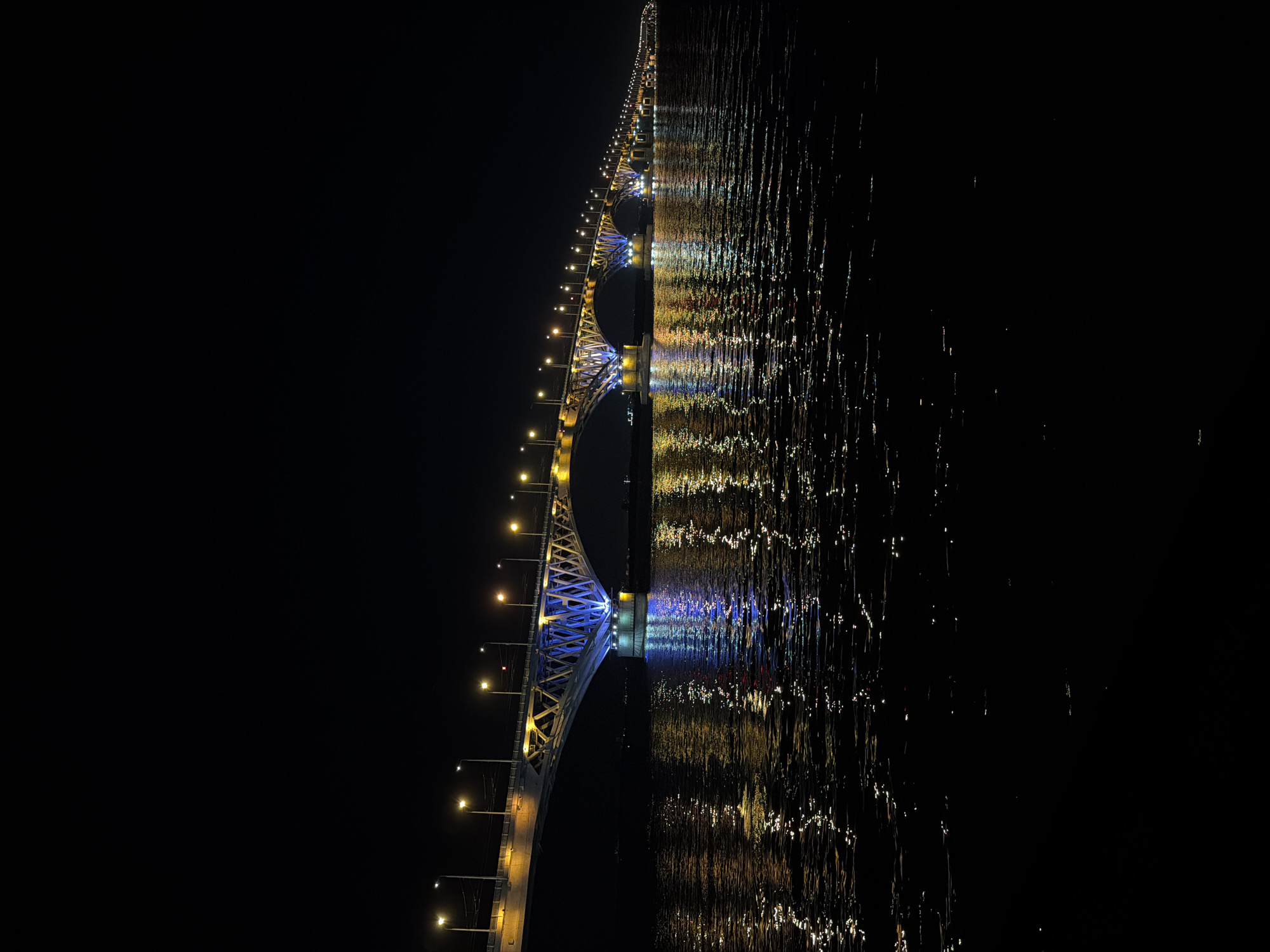 The image shows the Saratov Bridge, a large bridge spanning the Volga River, illuminated at night. The bridge, a significant piece of infrastructure in Saratov, Russia, is characterized by its long span and numerous lights. The lights on the bridge are a mix of yellow and blue, casting reflections on the dark, rippling water below. The water's surface is disturbed, creating a shimmering effect as it reflects the artificial illumination from the bridge. The sky above is completely black, indicating it is nighttime. There are no visible people or vehicles on the bridge or in the water. The scene is serene and highlights the architectural and lighting design of the bridge against the natural darkness of the night.