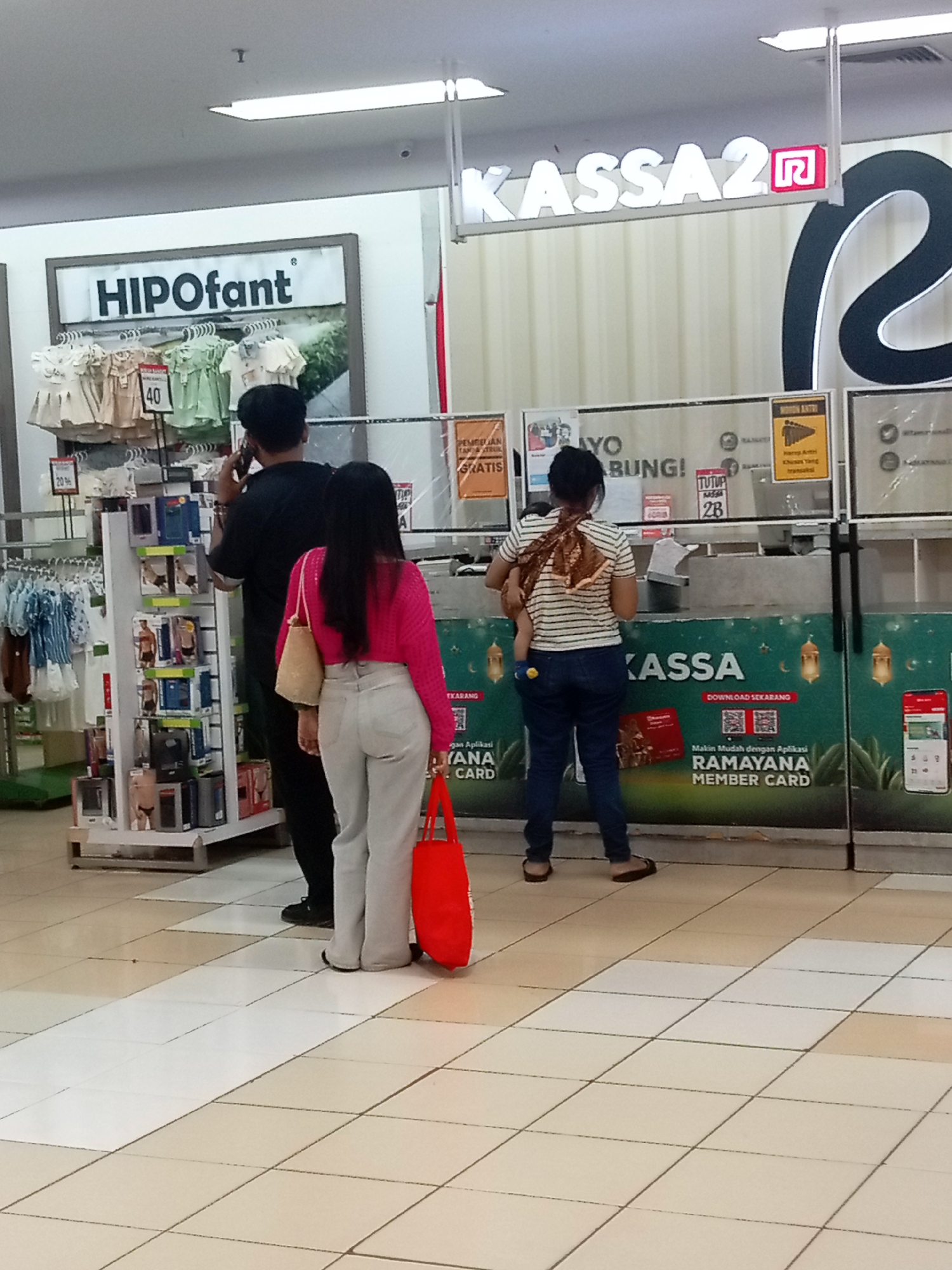 The image shows a retail environment, likely a department store in Batam, Indonesia. In the foreground, two individuals, a man and a woman, are standing near a checkout counter. The man, seen from behind, is wearing a black t-shirt and pants, and appears to be on his phone. The woman, also seen from behind, is wearing a bright pink long-sleeved top and light-colored pants. She is carrying a woven bag over her shoulder and a large red tote bag in her hand. She is looking towards the checkout counter. A third person, a woman, is visible in the background, further back in the queue, carrying a baby wrapped in a patterned cloth. The checkout counter has a prominent sign that reads "KASSA 2". Various promotional posters and displays are visible, including one that says "HIPOfant". The floor is tiled with a checkerboard pattern of cream and white squares. The lighting suggests an indoor commercial space.