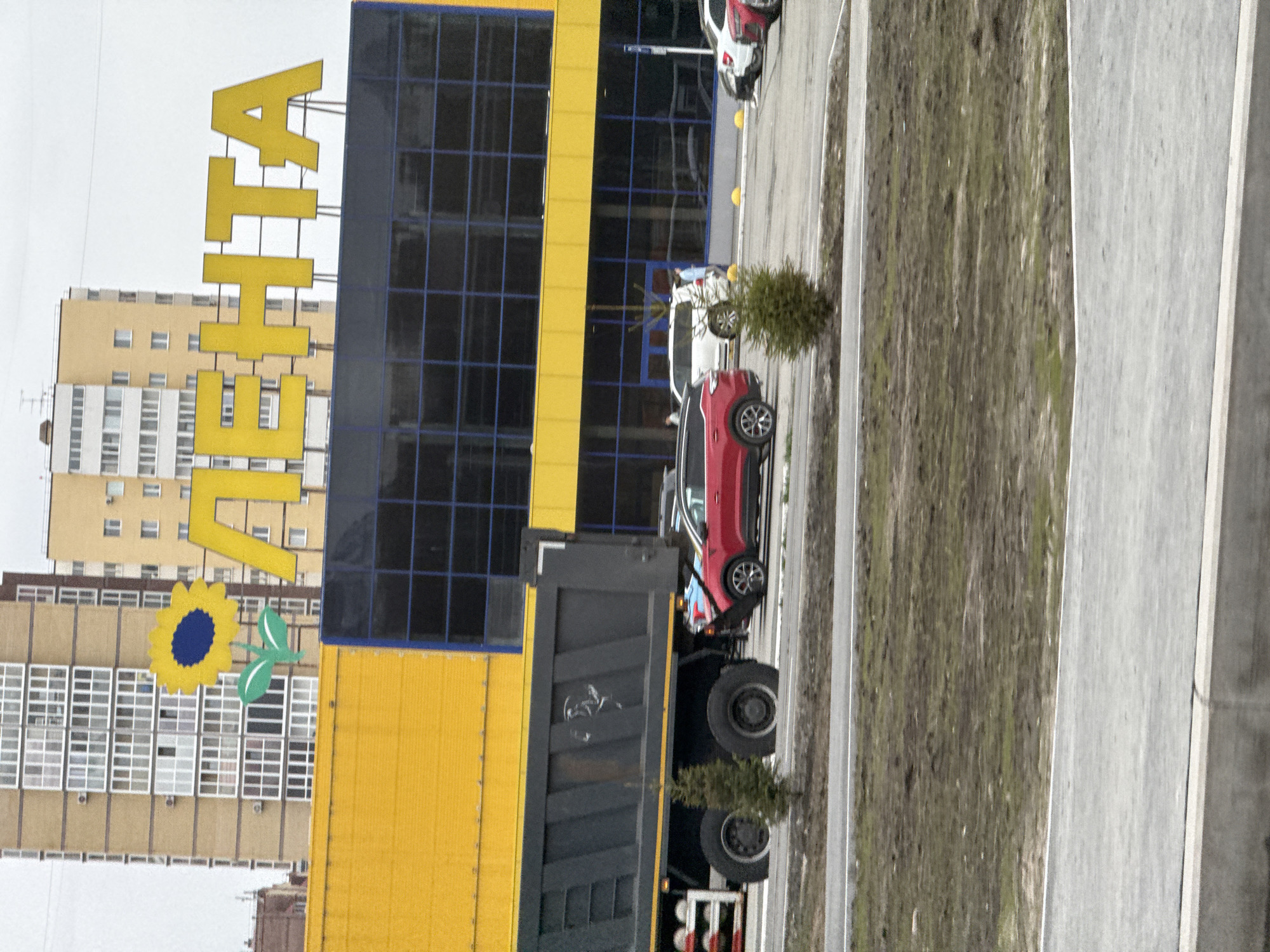 The image, taken in Tobolsk, Russia, captures an outdoor scene dominated by a large commercial building under an overcast sky. The building features a bright yellow facade and dark blue-tinted windows, arranged in a grid pattern. A prominent yellow sign spells out the Cyrillic word "ЛЕНТА" (LENTA) vertically along the building's side, identifying it as a Lenta hypermarket, a common Russian chain. Below the text, a large yellow sunflower with a blue center and green leaves, part of the Lenta logo, is affixed to the building.
In front of the building, a red SUV is parked, and a large dark grey dump truck is also present, partially obscuring the lower section of the building. Other vehicles, including a white car and another red car, are visible further in the paved parking area. The foreground includes a paved surface, strips of dry grass, and small evergreen bushes. In the background, several multi-story residential apartment buildings with beige and white exteriors are visible, characteristic of urban residential architecture. The overall impression is that of a bustling commercial and residential area during an overcast daytime.