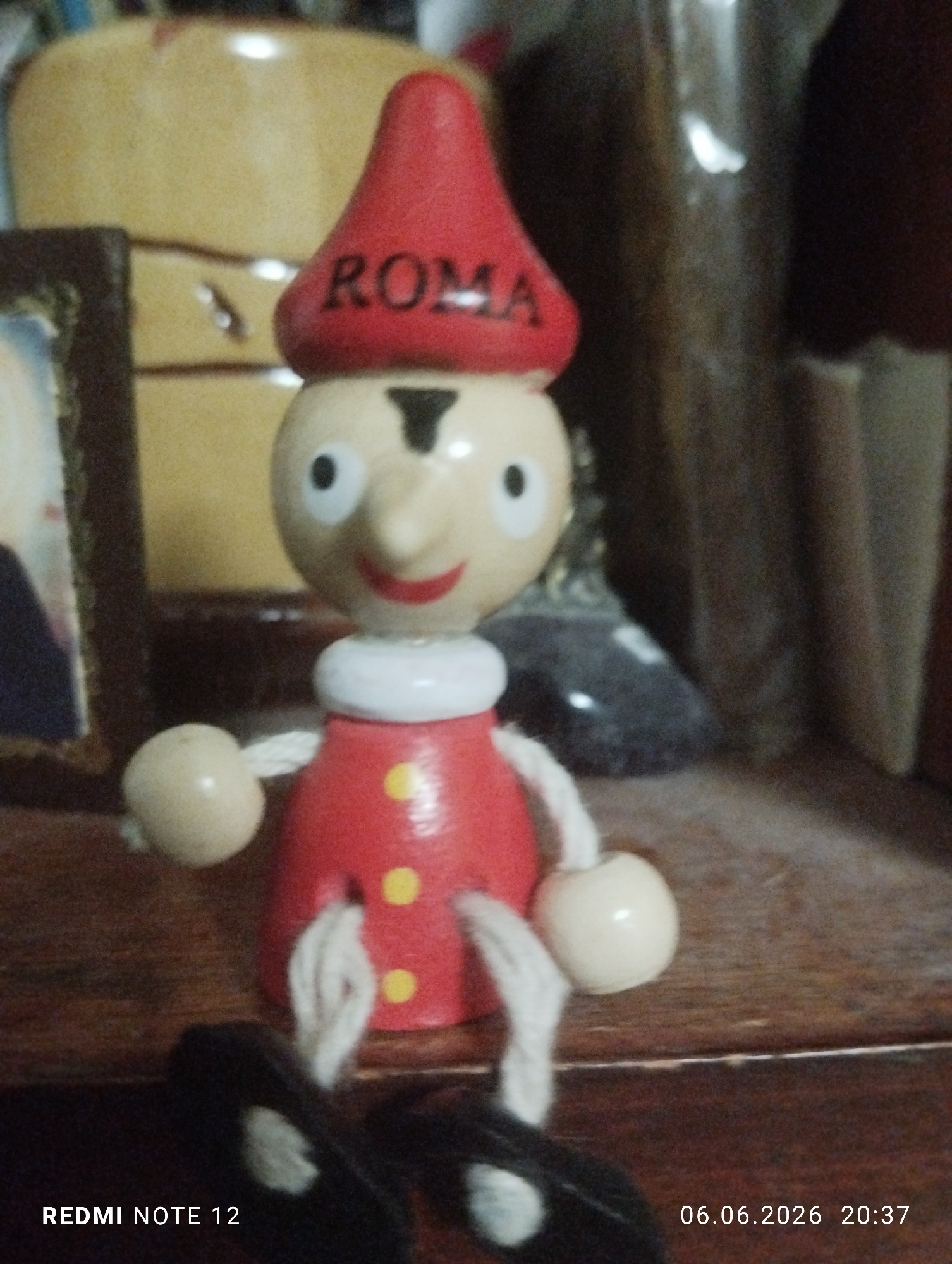 A wooden Pinocchio-style doll is centrally positioned on a dark wooden surface. The doll features a red conical hat with the word "ROMA" inscribed in black capital letters. Its head is light-colored, depicting black dot eyes, a red smile, a prominent nose, and a black T-shaped mark above the nose. A white collar separates the head from a red body adorned with three yellow circular dots vertically aligned on the front. Its limbs are composed of light-colored, spherical hands and feet connected by white string or cord. The doll's feet are partially obscured by a dark, soft object in the immediate foreground. In the background, to the left of the doll, a portion of a brown-framed item, possibly a picture, is visible. Behind the doll, a light tan or yellow object with dark markings is present. Further to the right and rear, additional dark, indistinct objects are discernible. The indoor environment suggests a shelf or table as the doll's resting place. The image was captured by a REDMI NOTE 12 device on June 6, 2026, at 20:37. The scene is located in the city of Burgas, Bulgaria.