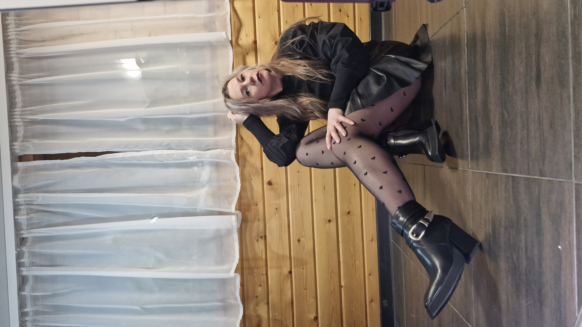 An indoor full-shot photograph, rotated on its side, features a woman posing within what appears to be a room in Otchor, Russia. The woman has long, light brown hair and fair skin. She is dressed in a black long-sleeved top, a black short skirt (possibly leather or faux leather), and black sheer tights adorned with small heart patterns. She completes her outfit with black high-heeled ankle boots featuring a buckle design.
The woman is depicted lying on her left side on a tiled floor, resting her head on her bent left arm. Her right arm is also bent, resting on her knee. She looks directly at the camera with a neutral expression.
The background consists of vertical wooden panelling behind her, and a window to her left covered by translucent white horizontal blinds or curtains, allowing soft natural light to enter. The floor is made of large, dark grey or brown rectangular tiles. The image captures a posed portrait in a residential or private setting during what appears to be daytime. There is no visible text in the image.