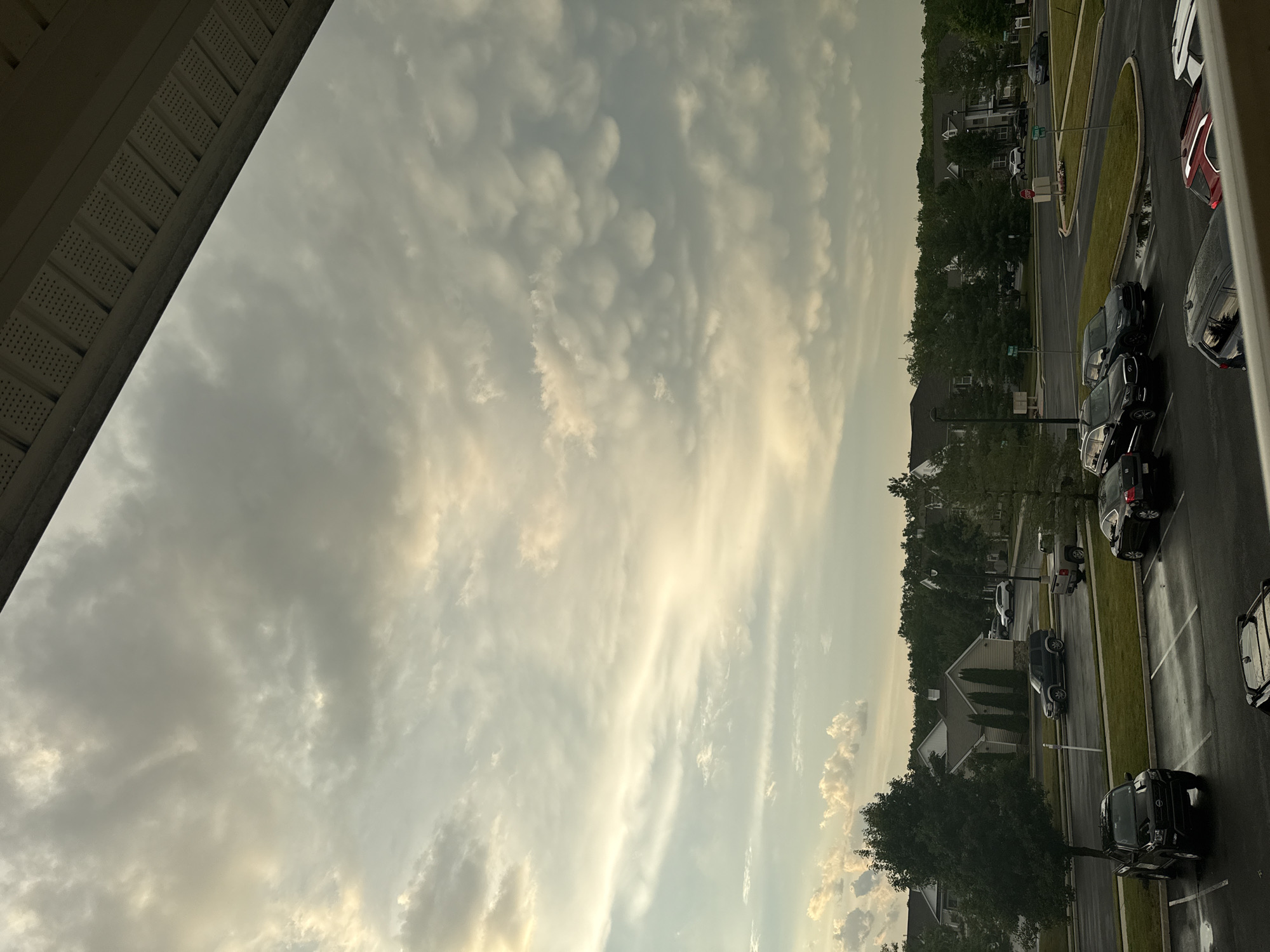 A wide-angle, eye-level view shows a suburban residential area under a dramatic, cloudy sky, possibly Allen Township, USA. The foreground is dominated by a parking lot with several vehicles, including a black pickup truck, a black SUV, a black sedan, a red sedan, and a white SUV. The asphalt is wet, indicating recent rain. Beyond the parking lot, a manicured lawn transitions to a street with more parked cars. Residential buildings with pitched roofs and landscaping are visible in the middle ground, surrounded by mature trees. The sky above is filled with a complex cloud formation, including mammatus clouds, illuminated by soft, diffused light, suggesting it is either early morning or late evening. A small part of a building's overhang or roof is visible in the upper portion of the frame, framing the scene from above.