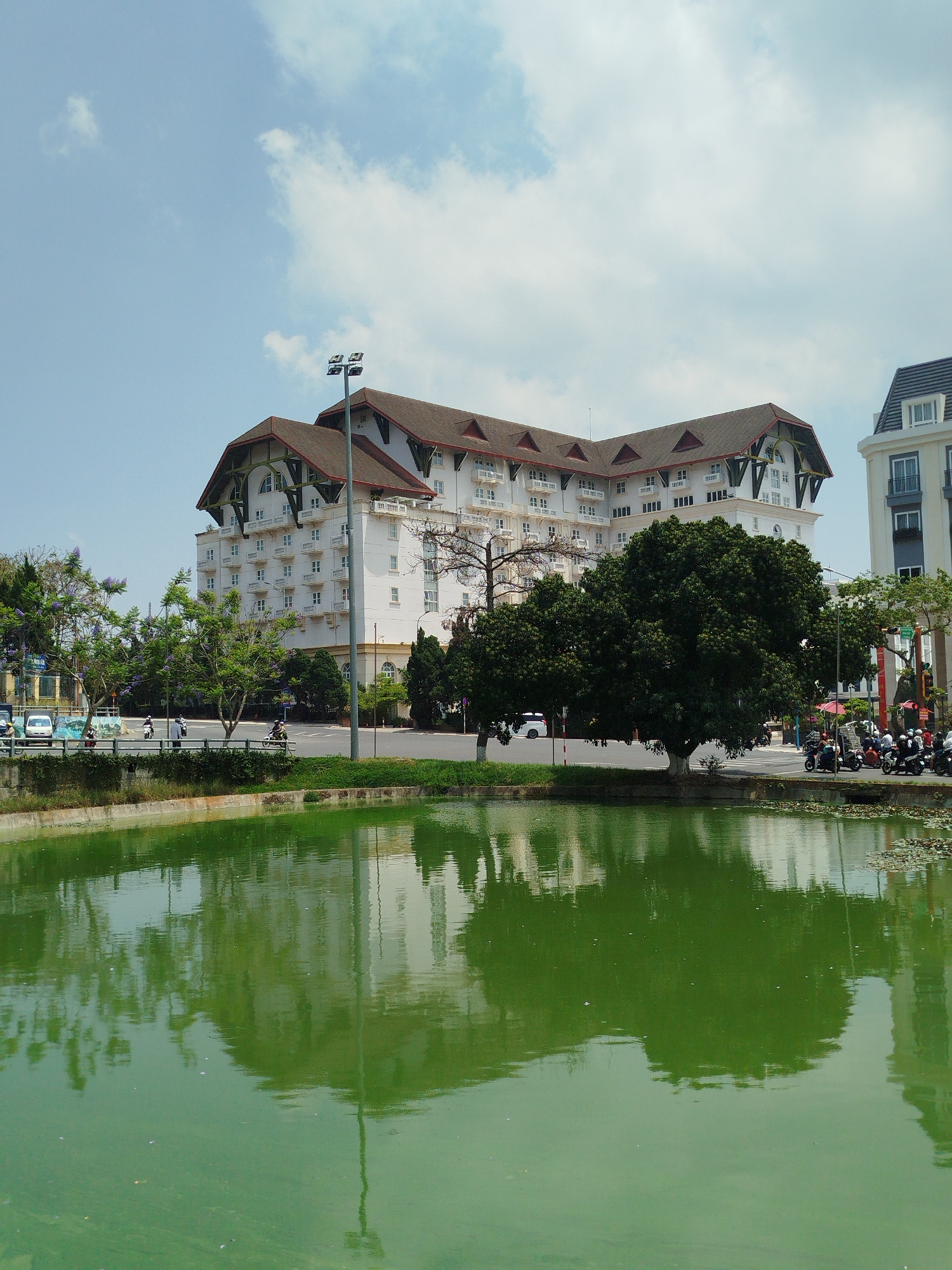 The image shows the Dalat Palace Heritage Hotel in Da Lat, Vietnam, reflected in a green pond. The hotel is a large, white building with a brown, multi-peaked roof, located behind the pond and separated from it by a road and landscaping. A tall lamppost stands near the hotel. Trees are positioned on the pond's edge. The water in the pond is a murky green. People and vehicles are on the road. Part of another building is on the right side of the image. The sky is blue with scattered white clouds.