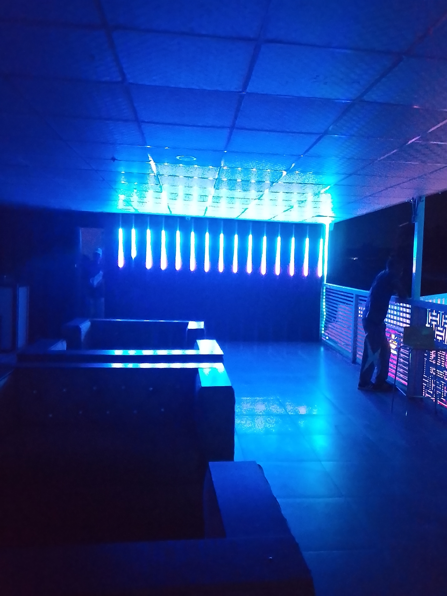 The image depicts an interior space, possibly a bar or lounge, illuminated predominantly by blue light. The ceiling has a grid-like pattern. A series of vertical light strips line the back wall, emitting a bright blue and green glow. Several dark, low seating arrangements are visible in the foreground. A person is standing near a railing decorated with illuminated geometric patterns. The location appears to be Narayi, Nigeria, based on the prompt.
