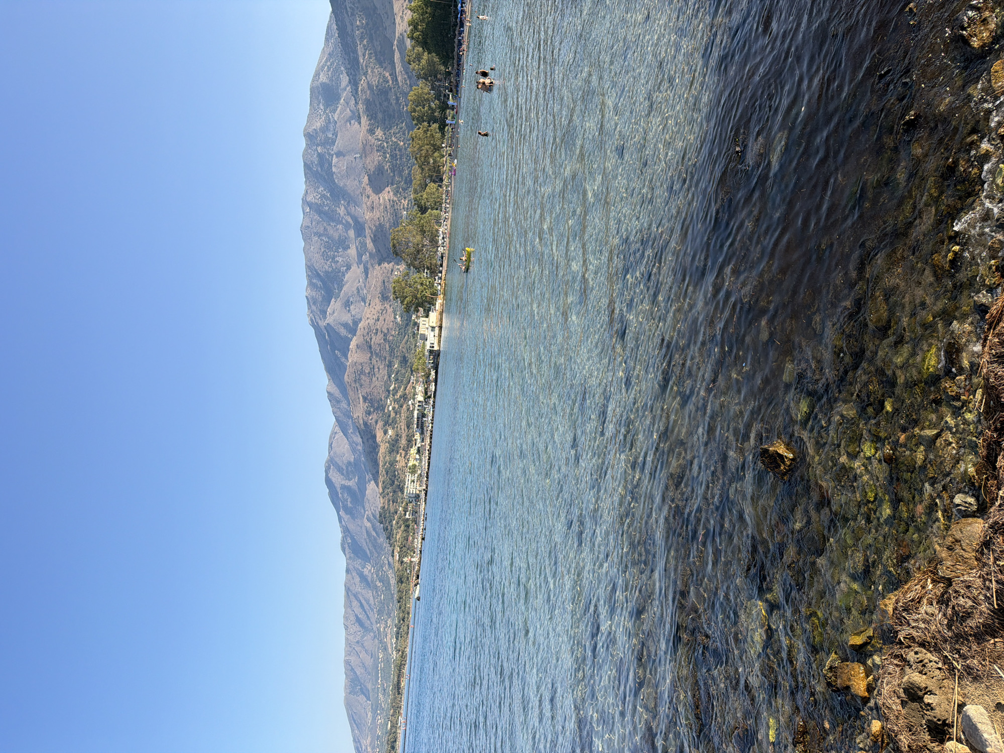 The image shows a clear, shallow sea with a rocky, seaweed-covered bottom near the shore. In the distance, people are swimming and wading in the water. Beyond the sea, a coastline is visible with buildings, trees, and a long stretch of beach lined with umbrellas and people. Further inland, rolling hills covered in vegetation rise to meet a large mountain range under a clear blue sky. The scene is consistent with a sunny day at a coastal resort. This specific view likely depicts the shoreline of Georgioupoli, Greece, looking towards the town and the surrounding mountains.