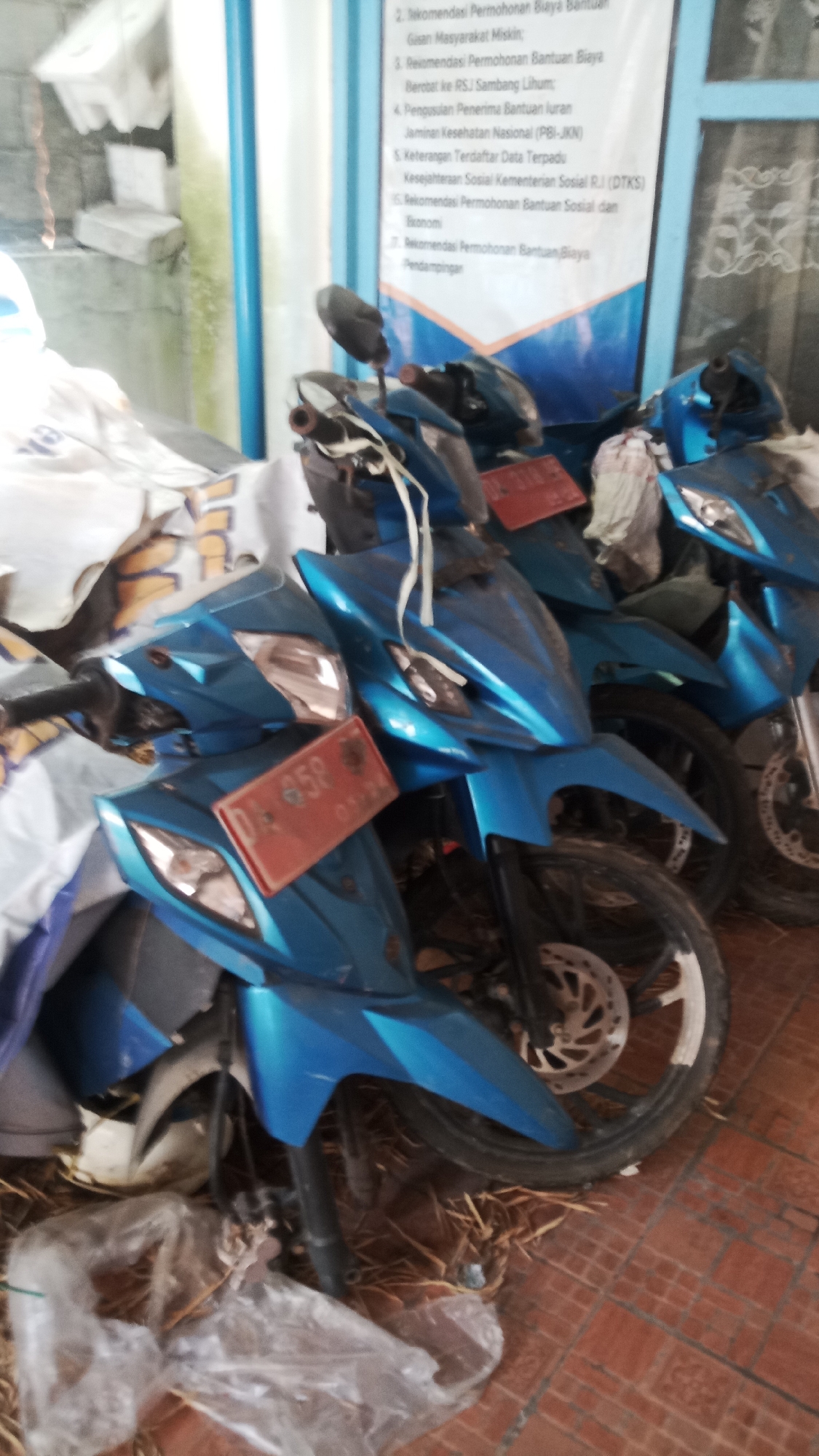 This image depicts a row of bright blue motorcycles parked indoors, likely in a storage area or a garage. The motorcycles are all the same model, characterized by their sporty design and vibrant color. One motorcycle in the foreground has a red license plate with white lettering that reads "D 658 01 24". The bikes are parked closely together, suggesting a limited space. Behind the motorcycles, a white banner with Indonesian text is visible, indicating a public or government-related context. The floor appears to be tiled with terracotta-colored tiles. Plastic bags and some debris are scattered on the floor around the motorcycles. The overall lighting suggests an indoor environment, possibly with natural light coming from an opening not fully visible in the frame. The scene is devoid of people.