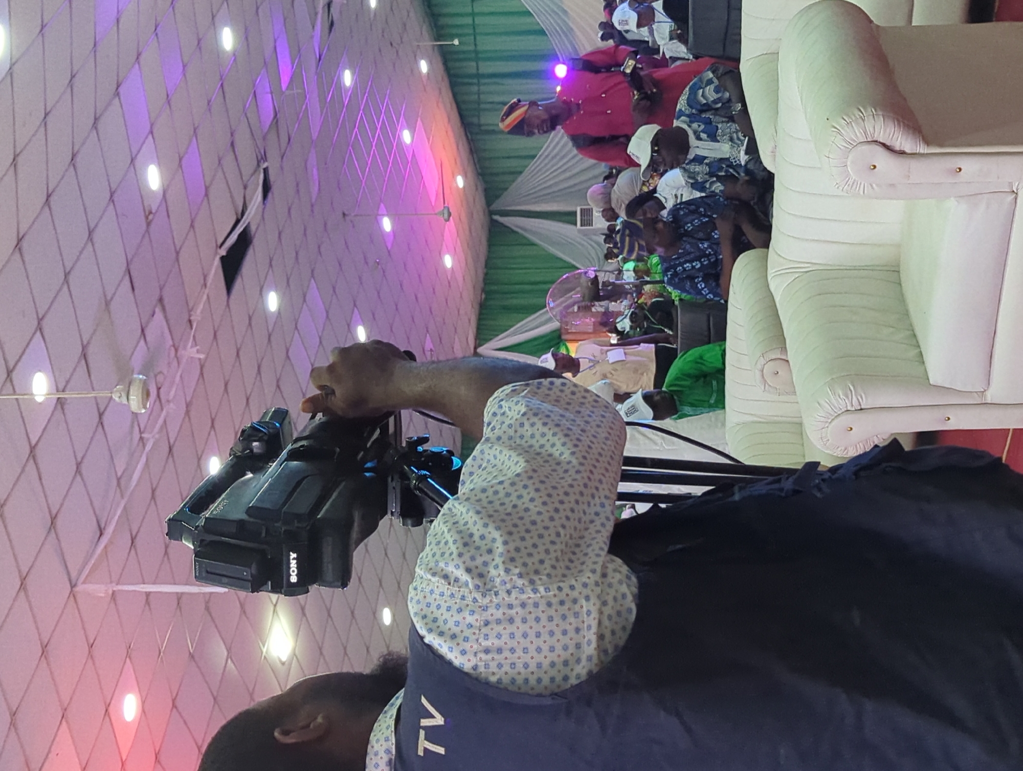 The image depicts a scene in Ungwan Liman, Nigeria, showing a cameraman operating a Sony video camera on a tripod. He is wearing a blue shirt with "TV" printed on it, as well as a patterned shirt underneath. The camera is focused on a group of people seated in the background, including one man wearing a red robe and hat. The setting appears to be an event or gathering, as indicated by the seating arrangement with white cushioned chairs and the presence of a decorated backdrop featuring green and white fabric. The ceiling is white with a diamond pattern and multiple recessed lights that cast a pinkish hue.