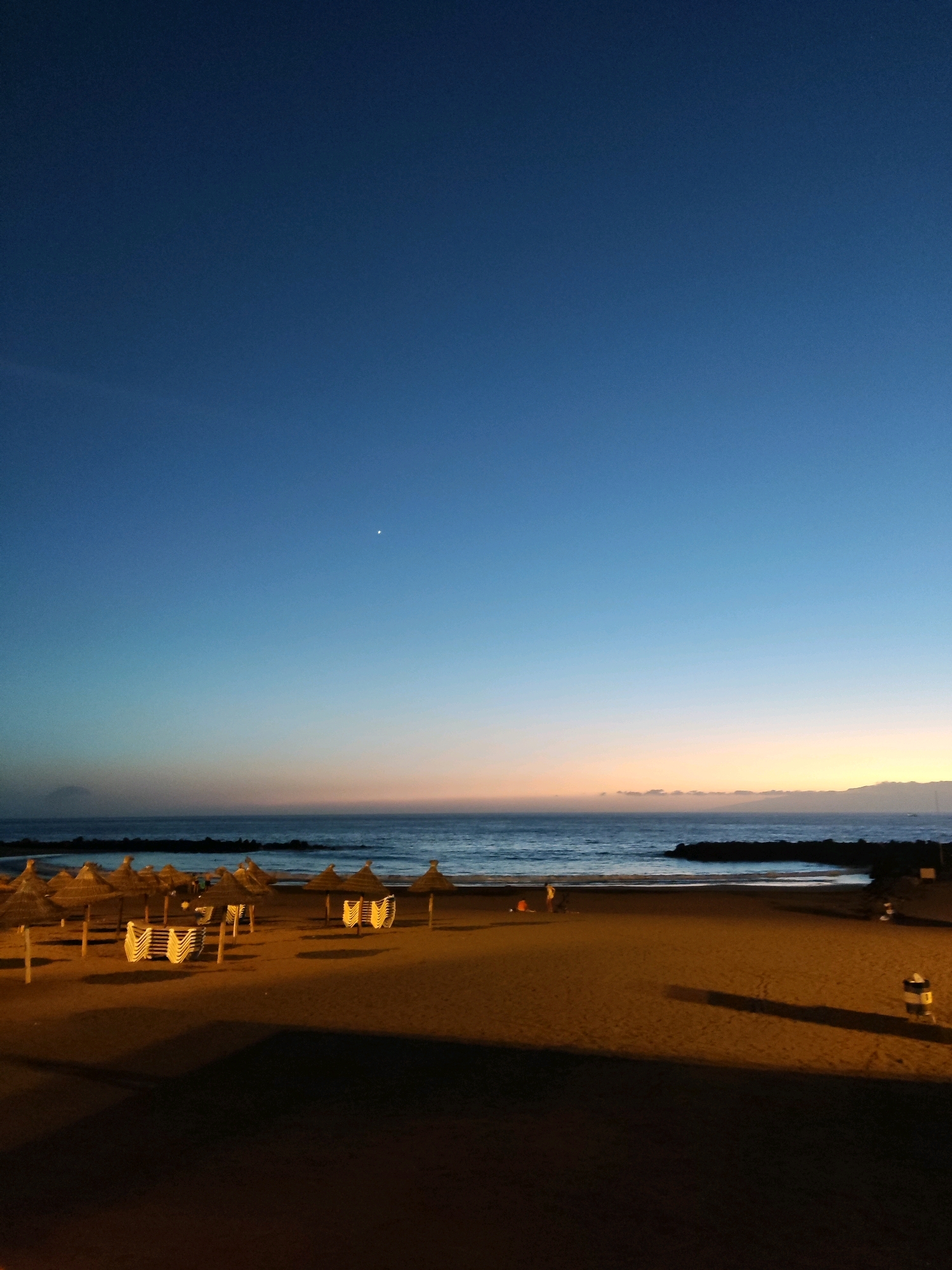 The image depicts a beach in Adeje, Spain, during twilight. The sky transitions from a deep blue overhead to soft hues of orange and pink near the horizon, where the sea meets the land. A single, bright star is visible in the upper portion of the sky.
The foreground of the image is a sandy beach, with rows of straw umbrellas and stacked beach chairs. The umbrellas are primarily clustered towards the left side of the frame, casting long shadows from artificial lighting on the beach. Some of these chairs are neatly stacked, while others are set up in small groups.
Further back, the sea is calm, with gentle waves lapping at the shore. A dark, rocky breakwater extends into the water on both the left and right sides of the frame, creating a sheltered area of the beach. Two small figures are visible on the wet sand near the water's edge on the right side of the image. Further to the right, a single trash receptacle is visible.
The overall atmosphere of the image is serene and tranquil, characteristic of a beach at dusk.