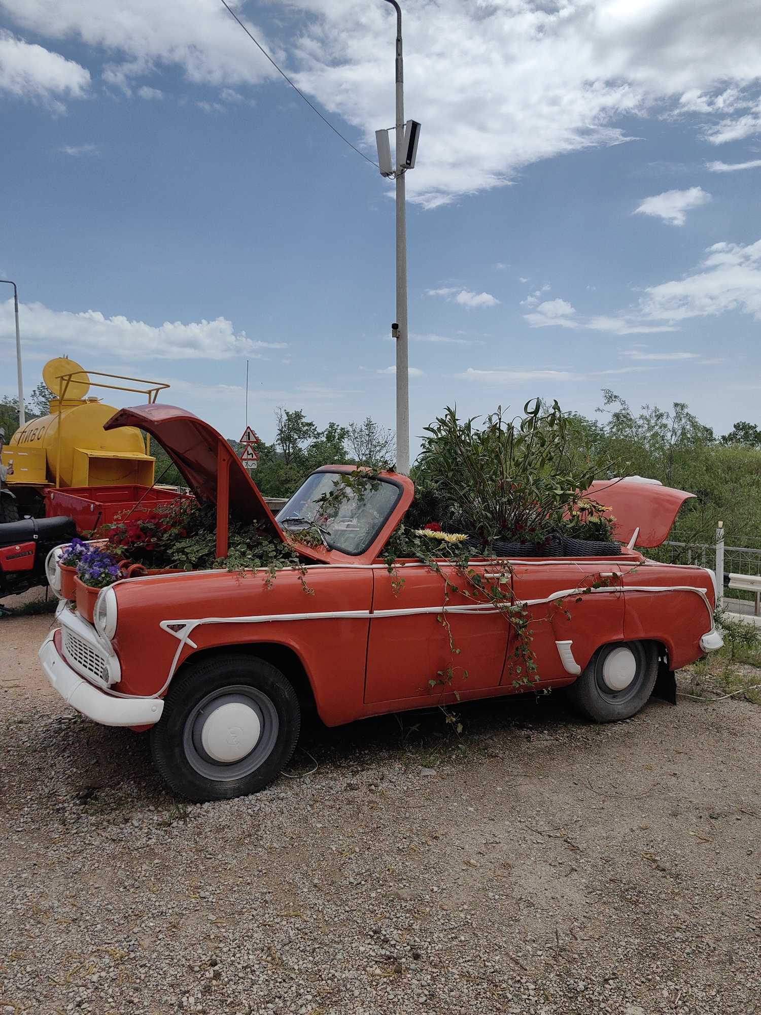 An orange Moskvitch car, repurposed as a flower bed, sits in Luchistoe, Ukraine. The hood and trunk are open, filled with various plants and flowers. The car is surrounded by a dirt and gravel ground. Behind the car, there's a yellow tanker and a white pole with lighting fixtures. A partly cloudy sky is visible above.