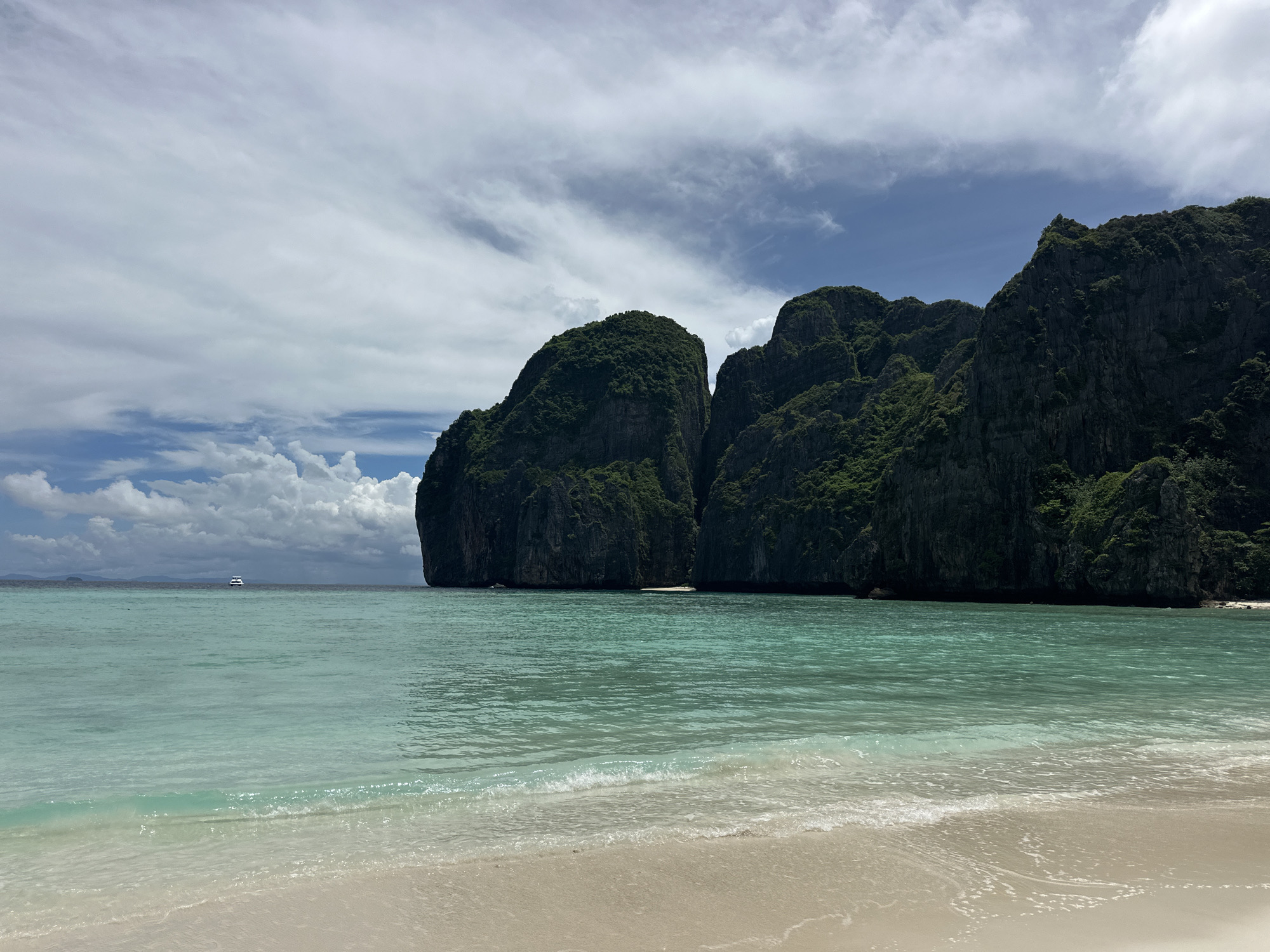 This is a photograph of Maya Bay, located on the island of Koh Phi Phi Leh, near Ao Nang, Thailand. The image depicts a tranquil beach scene with a body of turquoise water in the foreground, transitioning into a sandy shore. In the background, imposing limestone cliffs covered in lush green vegetation rise dramatically from the sea. The sky above is partly cloudy, with a few fluffy white clouds contrasting against a clear blue sky. A small white boat is visible in the distance on the calm water. The image captures the natural beauty of the tropical landscape.
