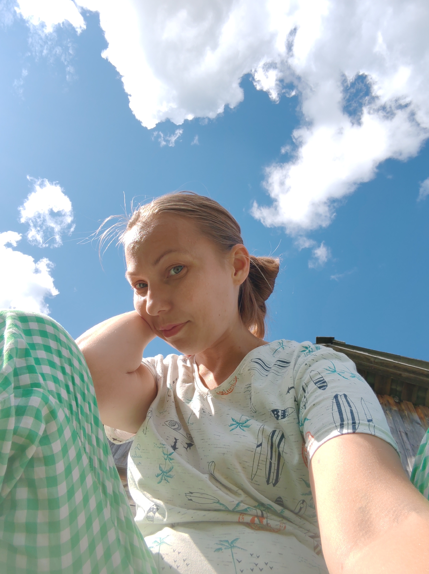 A woman with blonde hair pulled back in a ponytail is captured in a low-angle, outdoor selfie. She is wearing a white t-shirt with a pattern of surfboards and palm trees and green and white checkered pants. Her left arm is bent, and her hand is supporting her head as she looks towards the camera with a relaxed expression. The background is dominated by a bright blue sky with fluffy white clouds. To the right, the top of a wooden structure, possibly a building, is visible. The image appears to have been taken during daylight, with sunlight illuminating the scene.