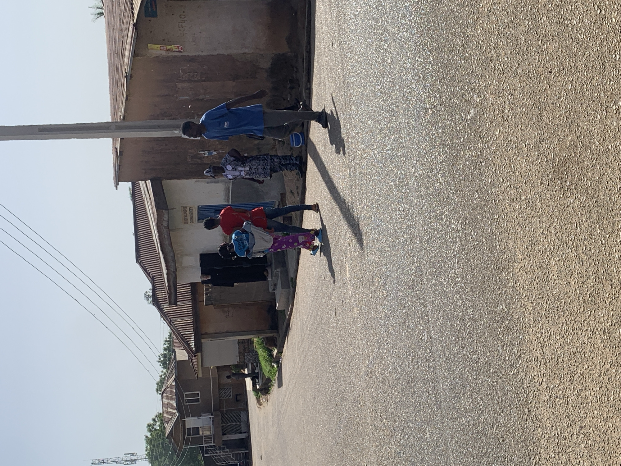 In Jos, Nigeria, a sunlit street scene shows a partially obscured building with a corrugated metal roof. An electrical pole stands near the building. A group of four individuals is walking alongside the building on the street. The first individual is a young man wearing a blue shirt and dark pants. The second is a woman in a patterned top and dark pants. The third is a person wearing a red shirt and jeans. The fourth is a child in a blue shirt and multi-colored patterned pants. The street surface is asphalt. Houses and trees are visible in the background. Electrical wires stretch above the street.