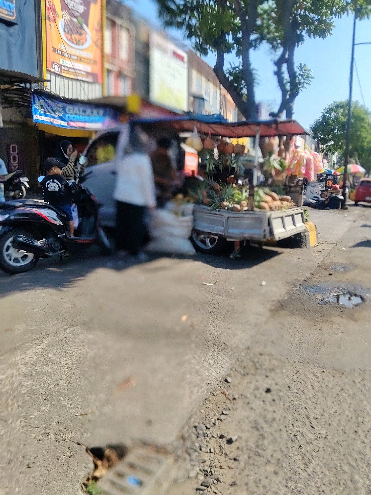 The image depicts a street scene in Cibatu, Indonesia, on a sunny day. In the foreground, a cracked asphalt road shows some puddles of water, suggesting recent rain or a spill. To the left, a black motorcycle is parked, with a young boy wearing a black t-shirt and a cap sitting on it. Behind the motorcycle, a person in a white shirt and black pants stands near a food stall.
Dominating the mid-ground is a small, mobile food cart laden with fruits, primarily pineapples and other items that are not clearly identifiable due to blurriness. The cart is covered by a red awning. The cart appears to be attached to a light-colored utility vehicle. In the background, several buildings line the street, some with signs, including one that reads "GALAXY". Trees with green foliage are visible, casting shadows on the road. Further down the street, other vehicles and stalls can be seen, including a red car and a stroller-like structure with a colorful umbrella. The overall impression is of a bustling street market or roadside commercial area.