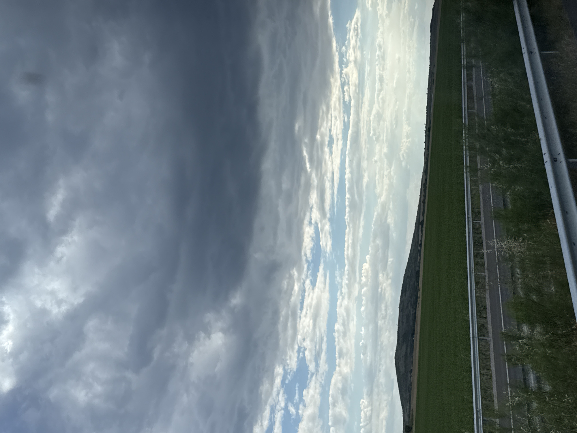 This is a horizontal, eye-level, medium shot of a rural landscape under a dramatic sky. The foreground features a highway with a silver guardrail running horizontally across the frame. Beyond the highway, a vast, green field stretches towards a rolling hill in the middle ground. The hill is covered in vegetation, and a small structure, possibly a tower, is visible on its crest. The background is dominated by a layered sky, with dark, heavy storm clouds occupying the upper portion of the frame, transitioning into lighter, wispy clouds and patches of blue sky towards the horizon. The lighting suggests it might be late afternoon or early evening. The scene depicts a natural environment with elements of human infrastructure. The location is identified as Polsko Padarevo, Bulgaria.