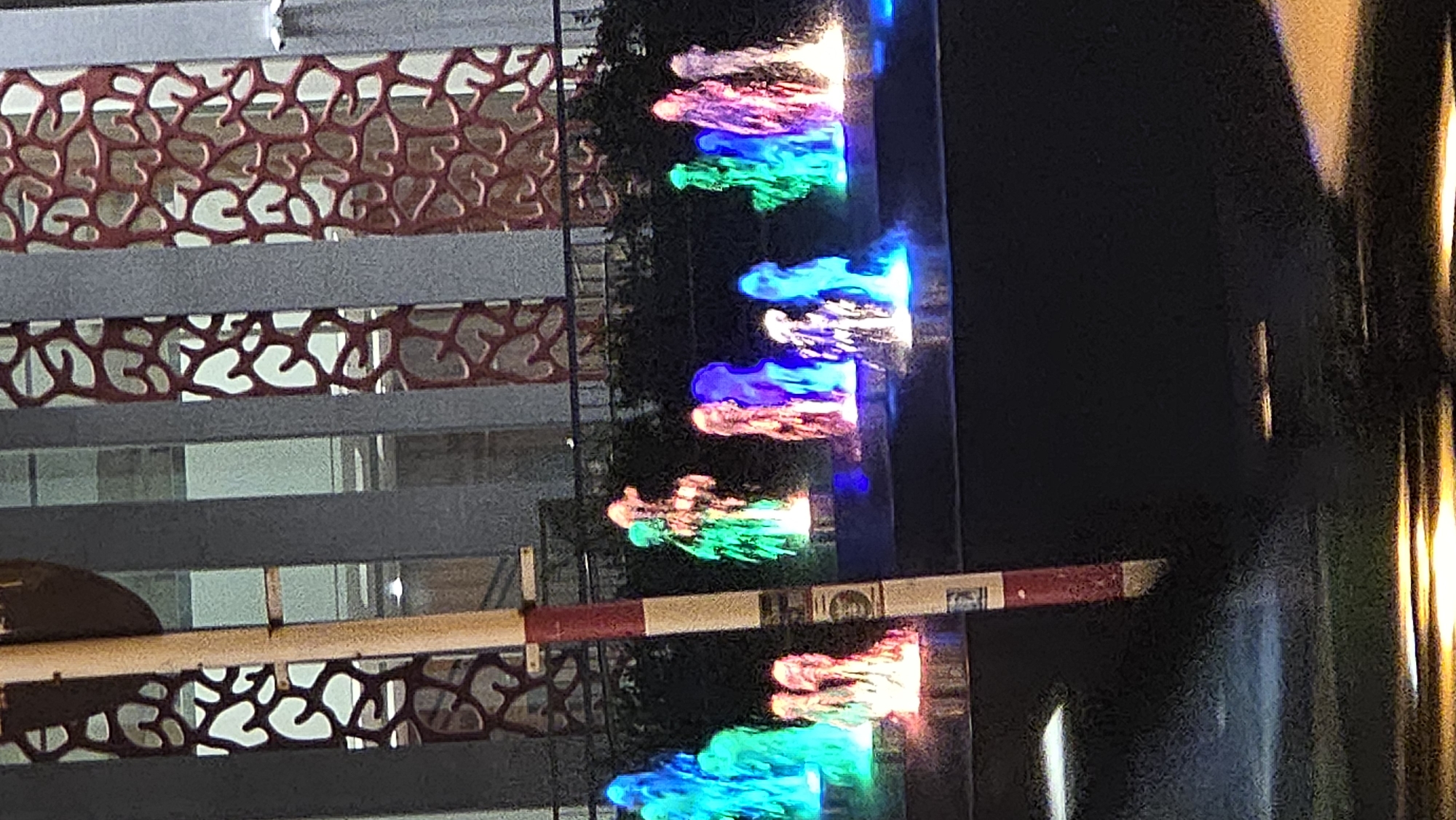 The image captures an outdoor night scene, likely in Nha Trang, Vietnam, featuring a decorative building facade, an access barrier, and illuminated water fountains.
On the left, a portion of a building or wall is visible, adorned with reddish-brown decorative panels that feature an intricate, organic, possibly coral-like pattern. These patterned sections are separated by horizontal grey bands, with hints of windows or reflective surfaces behind them.
A red and white barrier arm, commonly used for access control, extends horizontally across the lower-middle section of the frame. It displays a small, indistinct white rectangle with what appears to be an illegible logo or text.
The central and right parts of the image are dominated by multiple streams of water spraying upwards from fountains. These water jets are dramatically illuminated by various colored lights, including vibrant pink, blue, green, purple, and light blue hues, creating a lively and colorful display. Dark foliage, consistent with trees or bushes, forms a backdrop behind the fountains. On the far right, vertical elements emitting a warm yellowish light suggest additional architectural lighting or streetlights. The scene depicts an illuminated urban or resort area during nighttime, emphasizing decorative lighting and water features.