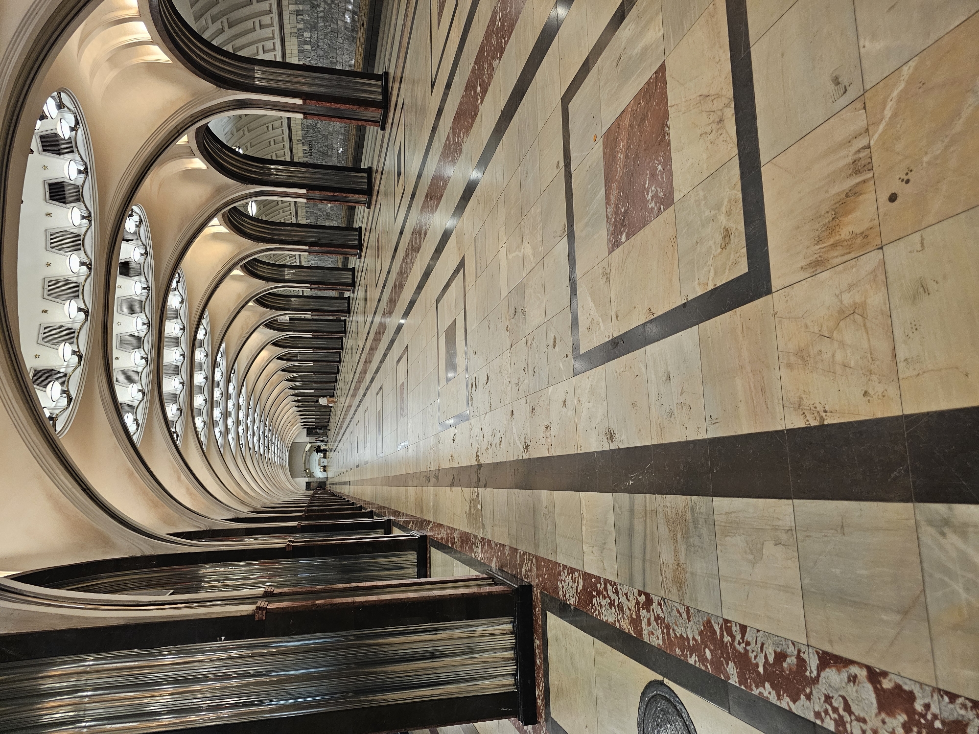 This is a wide-angle, eye-level shot of a grand, cavernous metro station concourse in Moscow, Russia. The architecture features a repeating pattern of high, arched ceilings supported by numerous large, dark columns, creating a sense of depth and perspective that draws the viewer's eye down the long passageway. The ceiling is adorned with numerous symmetrical, circular light fixtures. The floor is paved with large, rectangular tiles of varying light colors, bordered by darker stripes and framed by rectangular insets of reddish-brown marble. There are a few distant, silhouetted figures walking in the concourse, suggesting a public space in use. The overall impression is one of grandeur, symmetry, and architectural significance.