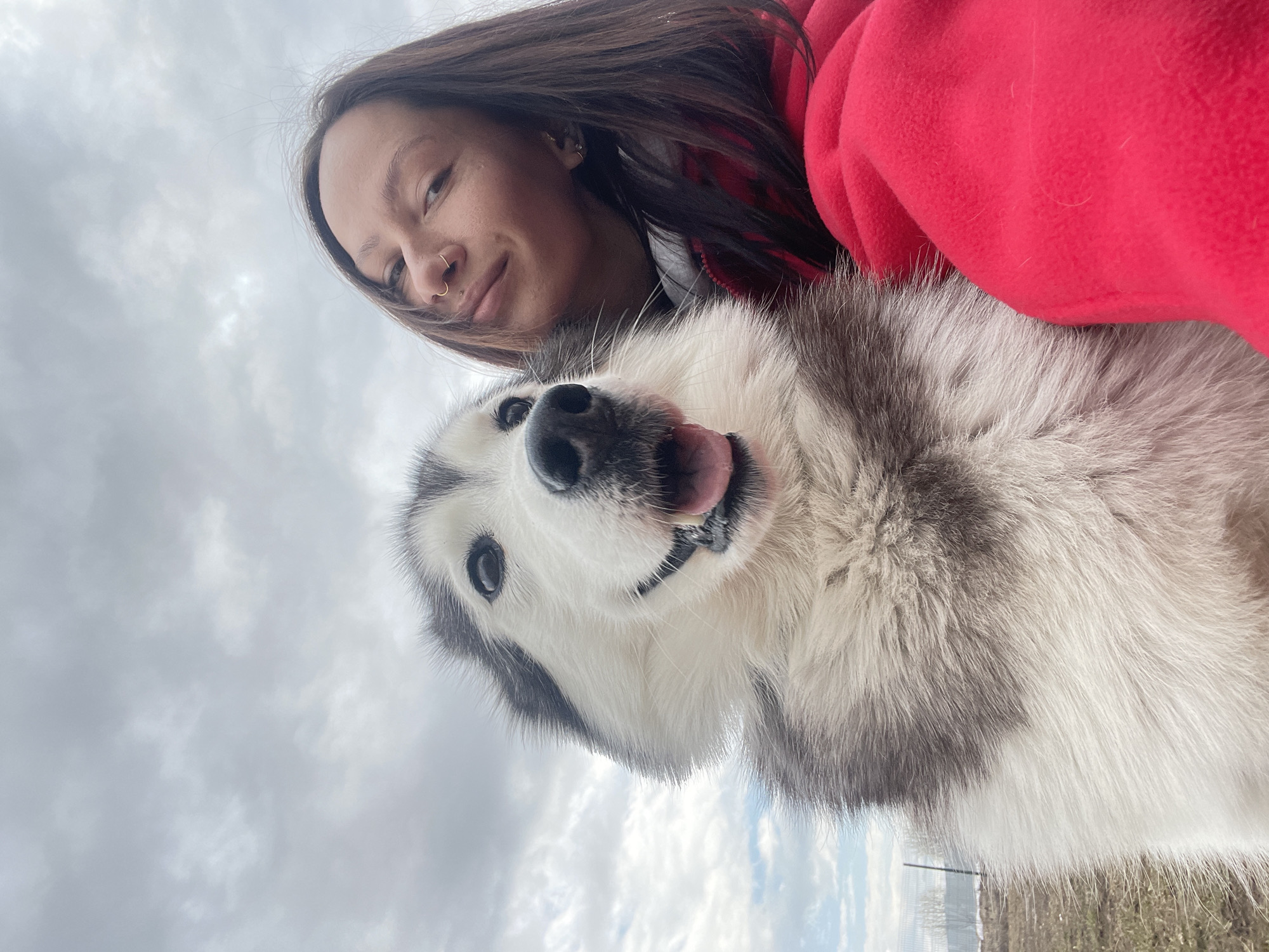 A close-up, upward-angled portrait features a young woman and a fluffy husky dog, captured in what appears to be a selfie. The image is oriented vertically, with the subjects appearing tilted against the sky.
The woman, positioned on the left and slightly behind the dog, has long dark brown hair, fair skin, and a gentle smile. She wears a golden nose ring on her right nostril and a small stud above her upper lip. Her attire is a vibrant red fleece jacket.
The husky dog occupies the central and right portions of the frame, with its face prominent. Its fur is a mix of white, grey, and black, typical of the breed. The dog's mouth is slightly open, and its tongue is extended, conveying an expression of happiness or exertion. Both the woman and the dog are looking towards the camera.
The background is dominated by an overcast sky filled with light grey clouds, suggesting an outdoor setting during daytime. Minimal ground is visible at the very bottom edge of the image (if rotated correctly), showing faint hints of dry vegetation and possibly distant structures or fences. The scene captures a warm moment between the woman and her pet, likely in an open area of Izhevsk, Russia. There is no visible text in the image.