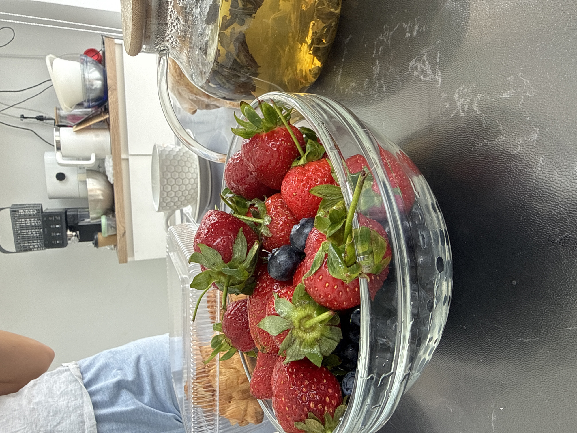 A close-up, rotated image shows a glass bowl filled with strawberries and blueberries. The bowl sits on a dark surface, possibly a countertop. Behind the bowl, a glass teapot filled with a yellow liquid (likely tea) is partially visible. Further to the left, a kitchen counter with various appliances and dishes is present. In the lower left corner, a person wearing light blue clothing is partially visible. The location is Olginskaya, Russia.