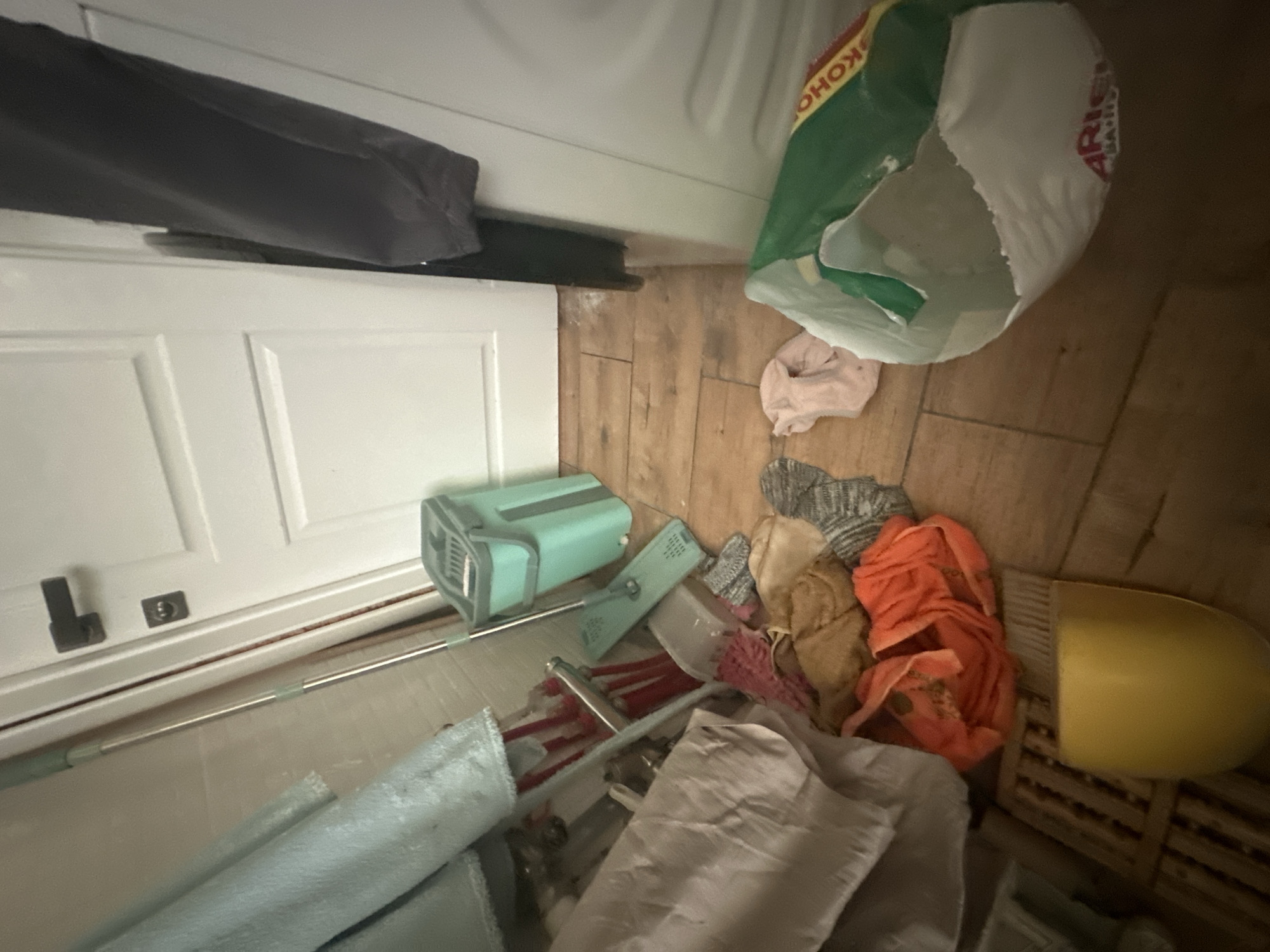 The scene is an interior space with a closed white door in the lower-left and a white cabinet or appliance along the top edge. A gray cloth lies atop the door. On the right, there is a partially visible green and white plastic bag. The floor is covered with light-brown wooden tiles, and several cleaning items are strewn about. These include a light-blue mop with a metallic handle, a green cleaning device with a gray handle, an orange towel, a yellow dustpan, and assorted cloths in various colors. The location is unknown.
