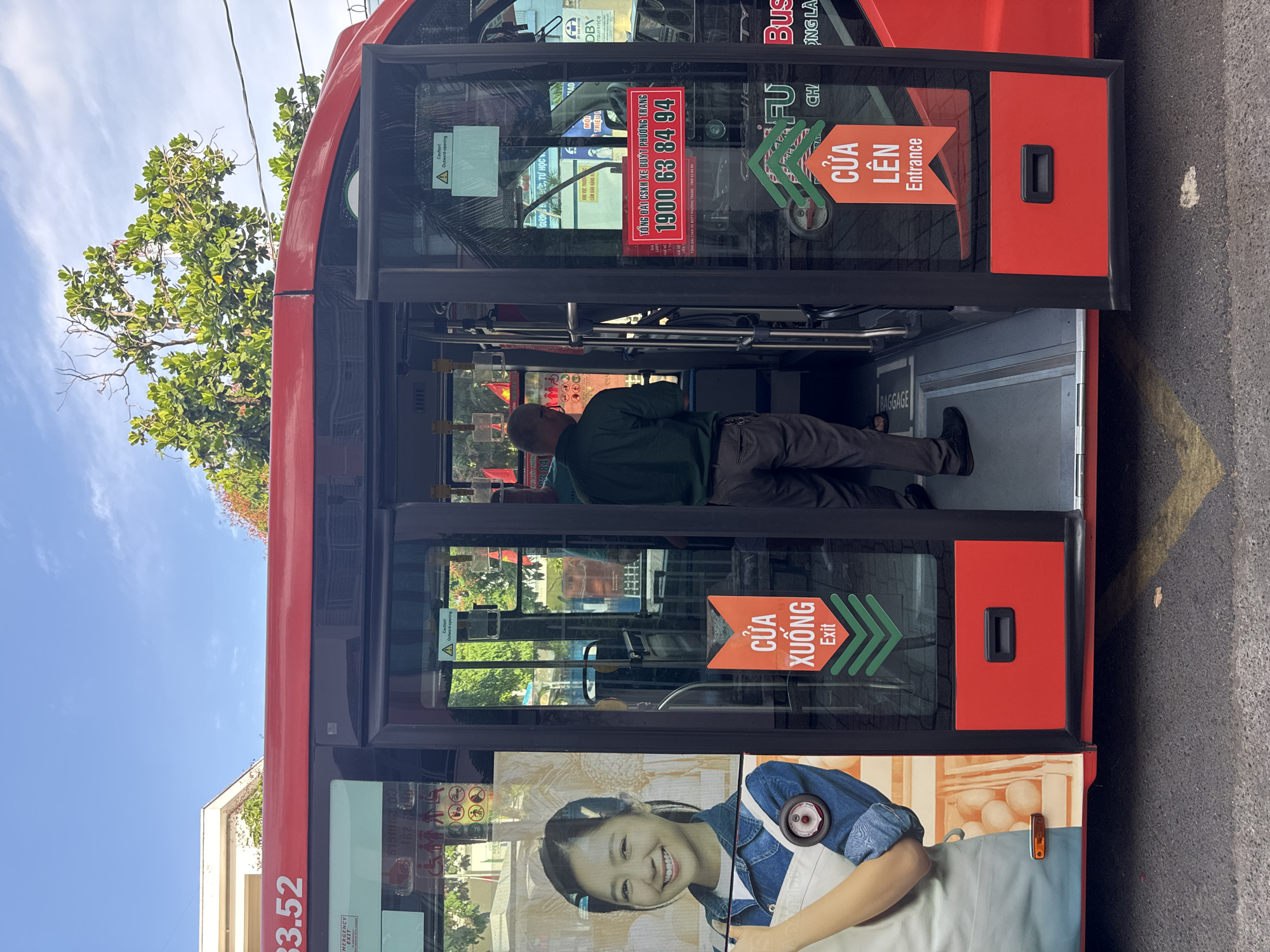 The image shows the open doors of a red public bus in Nha Trang Ward, Vietnam. On the left side of the bus, a large advertisement features a smiling woman wearing a denim shirt and an apron, with a basket of eggs visible. Two signs are prominently displayed on the bus doors. One, a red sign with green accents, indicates "CỬA XUỐNG Exit." Below it, another red sign with green accents reads "CỬA LÊN Entrance." A man in a green shirt and dark pants is walking into the bus through the open doors. The bus appears to be stopped at a bus stop, as indicated by the road and sidewalk. The sky is blue with scattered clouds, and green trees are visible in the background. The overall scene depicts a typical day on public transportation in Vietnam.