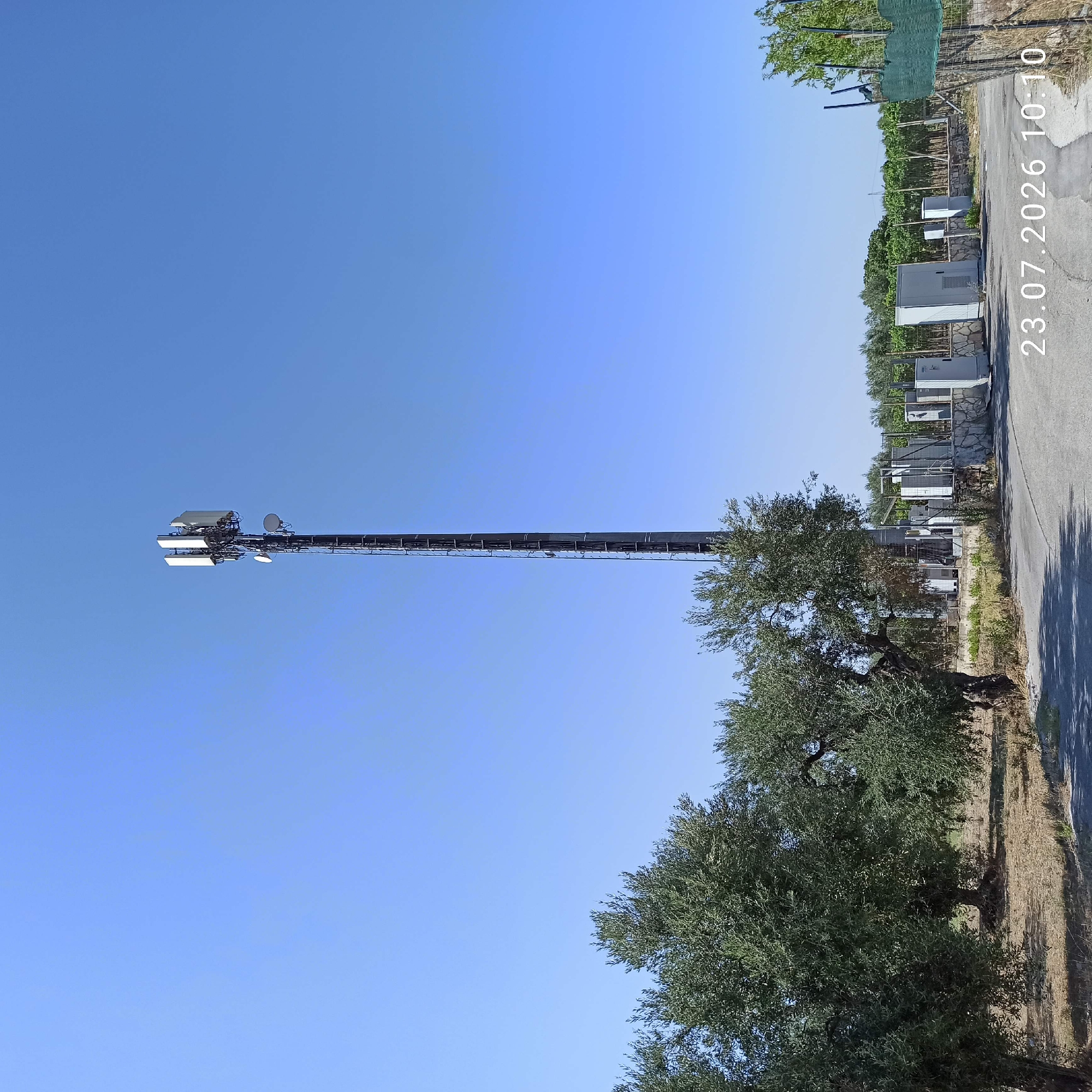 An outdoor, eye-level, full shot captures a telecommunications tower situated in Bitonto, Italy, under a clear, bright blue sky. The tower, a tall, dark metal lattice structure, is adorned with various antennas and equipment at its apex. It stands prominently in a rural landscape characterized by dry, light-brown earth and sparse vegetation. To the left of the tower, several mature olive trees with gnarled trunks and dense green foliage are visible. In the foreground, a grey asphalt road curves around the scene. At the base of the tower, a fenced-off area contains several white utility boxes and structures. The date and time "23.07.2026 10:10" are superimposed on the bottom right of the image.