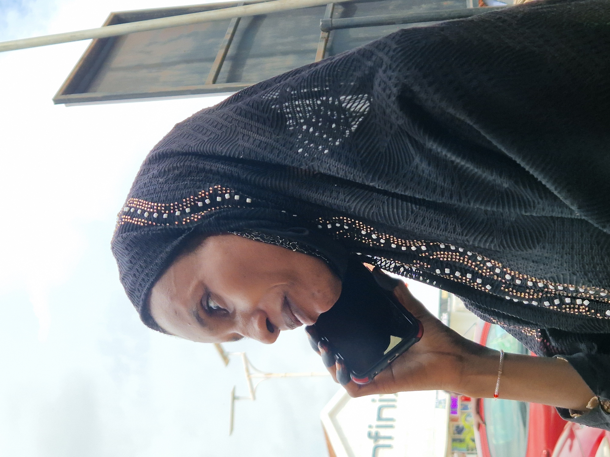A woman wearing a black hijab with intricate beaded detailing is speaking on a black smartphone. She is looking downwards and to her left, with a slight smile on her face. Her left arm is raised, holding the phone to her ear. A thin bracelet with red and white beads is visible on her wrist. The background suggests an outdoor setting with a bright, possibly cloudy sky. Partially visible in the background are the edges of a metal structure and a sign with the text "nfin". The image is oriented sideways.