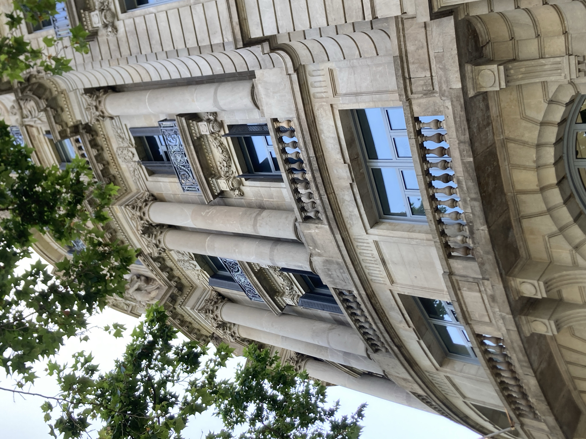 The image shows a low-angle, full-shot view of the exterior of a grand, ornate building in Barcelona, Spain. The architecture features classical elements such as large, fluted columns, intricate stone carvings, and decorative balconies with balustrades. The building's facade is made of light-colored stone, with multiple stories visible, each adorned with windows and architectural details. Some windows are open, revealing dark shutters. Green foliage from trees drapes down from the top of the frame, partially obscuring the upper sections of the building. The sky is overcast and light, providing soft illumination. The building appears to be a historic structure, possibly a public building or a palatial residence.
