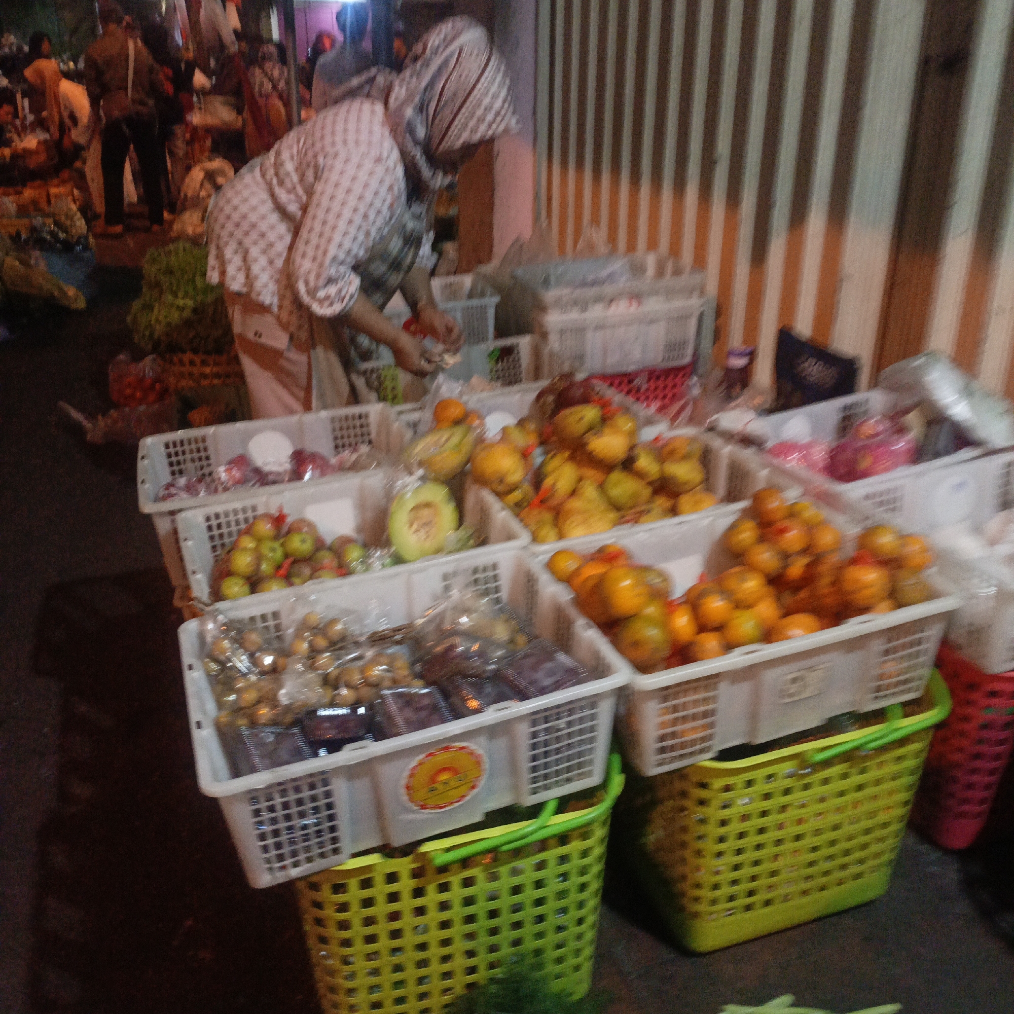 The image depicts a street market scene in Salatiga, Indonesia, likely at night or in low light conditions, as indicated by the artificial lighting and shadows. A woman, partially visible and wearing a head covering and a checkered shirt, is attending to her stall. She is seen reaching into a white plastic basket filled with fruits. The stall is laden with various fruits displayed in white plastic crates and baskets. Prominently featured are green apples, yellow citrus fruits, and possibly grapes or similar small fruits, some of which are packaged in transparent plastic. A large green plastic basket with a checkered pattern is positioned below one of the fruit crates, and a red basket is partially visible to the right. In the background, striped vertical elements, possibly corrugated metal or bamboo siding, form a wall or structure. Other market-goers and stalls are visible in the blurred background, suggesting a busy market environment. The overall impression is one of a lively, albeit somewhat chaotic, marketplace.