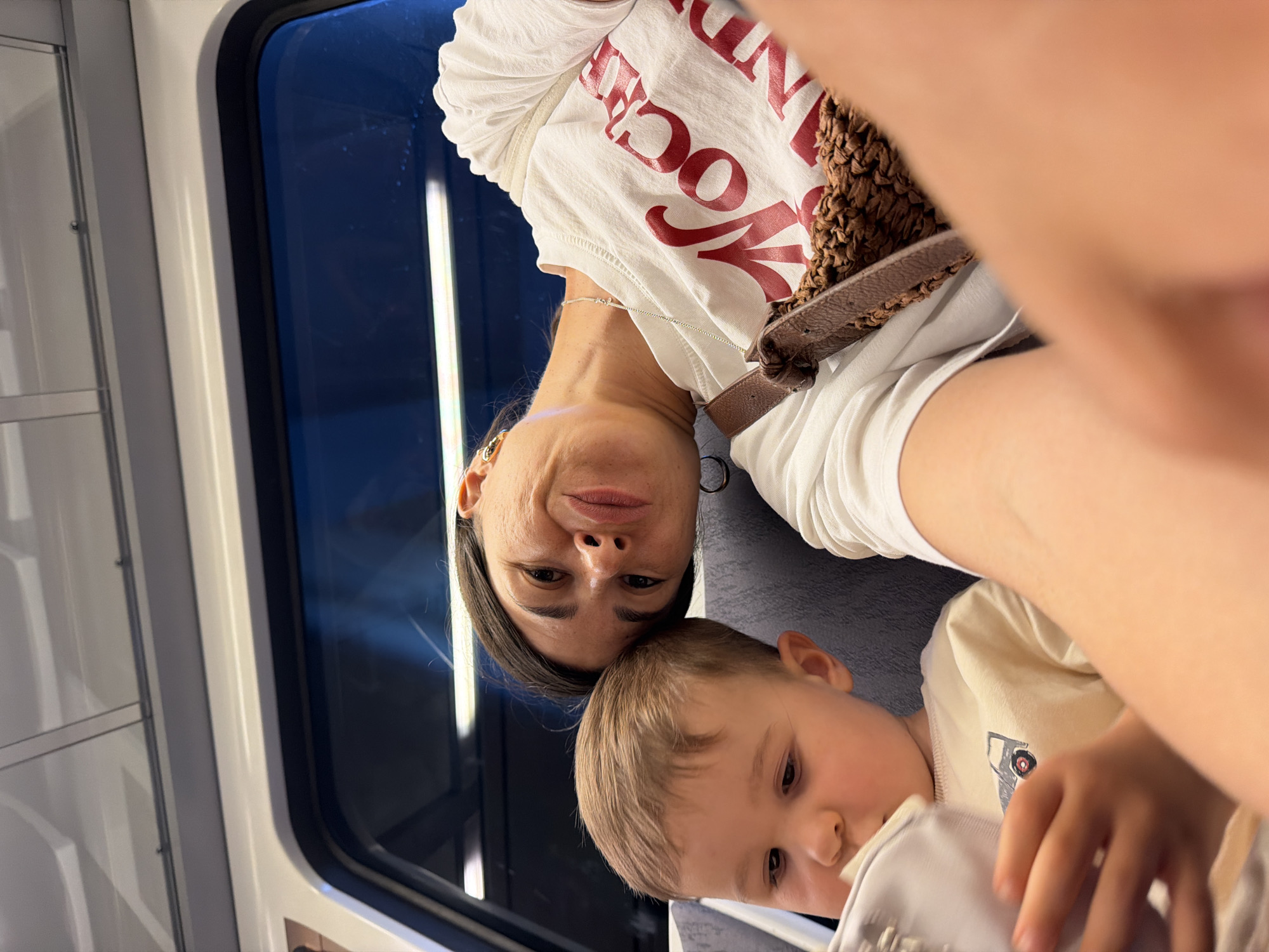 A woman and a young child are seated on a train, with the woman resting her head on the child's. The woman is looking at the camera and smiling, while the child is looking down at something in their hands. The woman is wearing a white t-shirt with red text on it, and the child is wearing a light-colored t-shirt with a picture of a truck. A brown woven bag is slung across the woman's chest. The train car appears to be modern, with a large window showing a dark, blurry view of the outside. The lighting suggests it is either evening or a dimly lit interior. There is no indication this photo was taken in Barcelona, Spain.