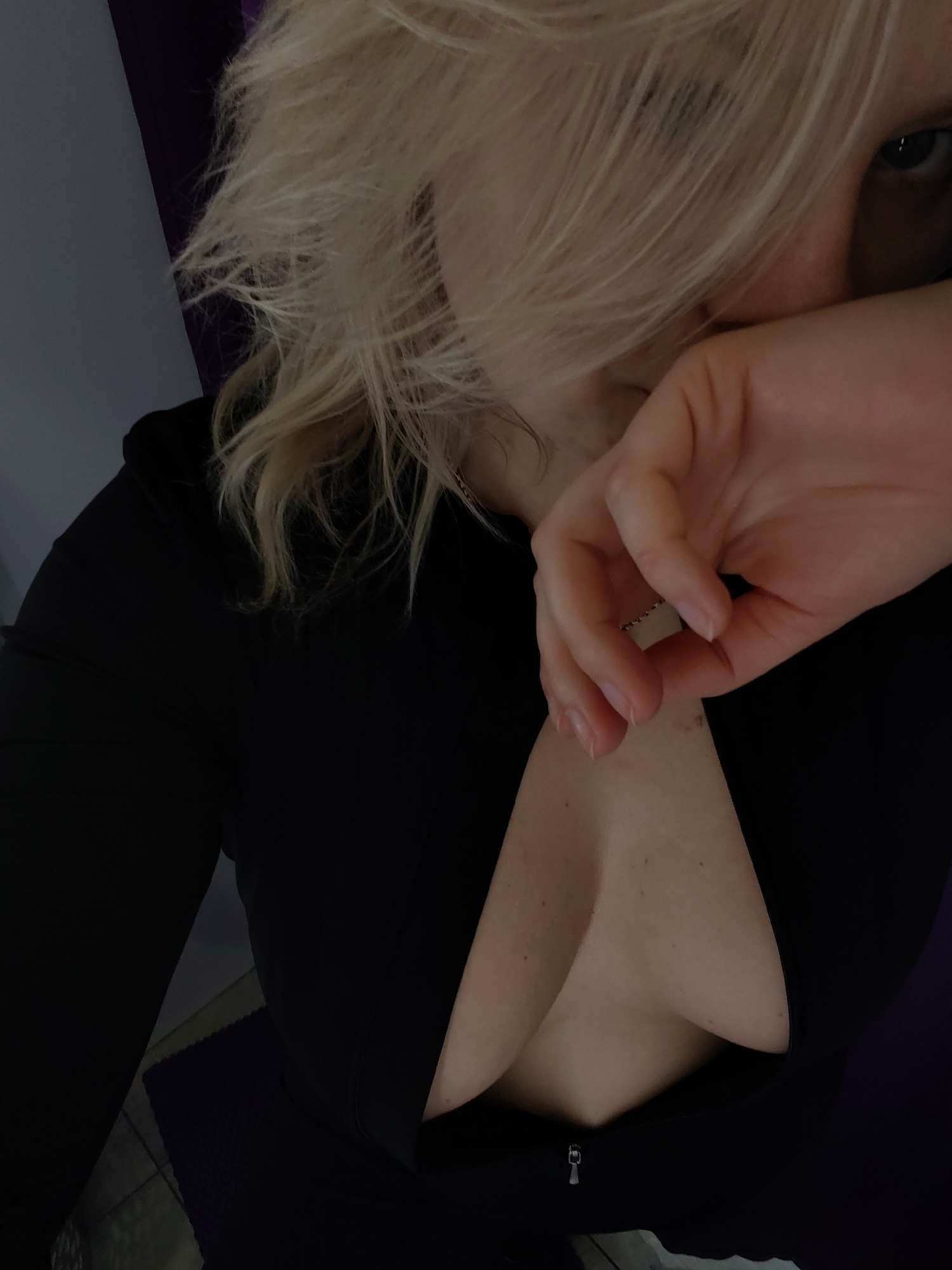 The image is a close-up selfie of a woman with blonde hair, taken from a high angle. She is wearing a black, low-cut top. Her right hand is raised to her face, partially obscuring it, and she is looking at the camera. The background is dark and indistinct, with hints of purple and gray. There is no information in the image to indicate the location in the city of Izhevsk, Russia.