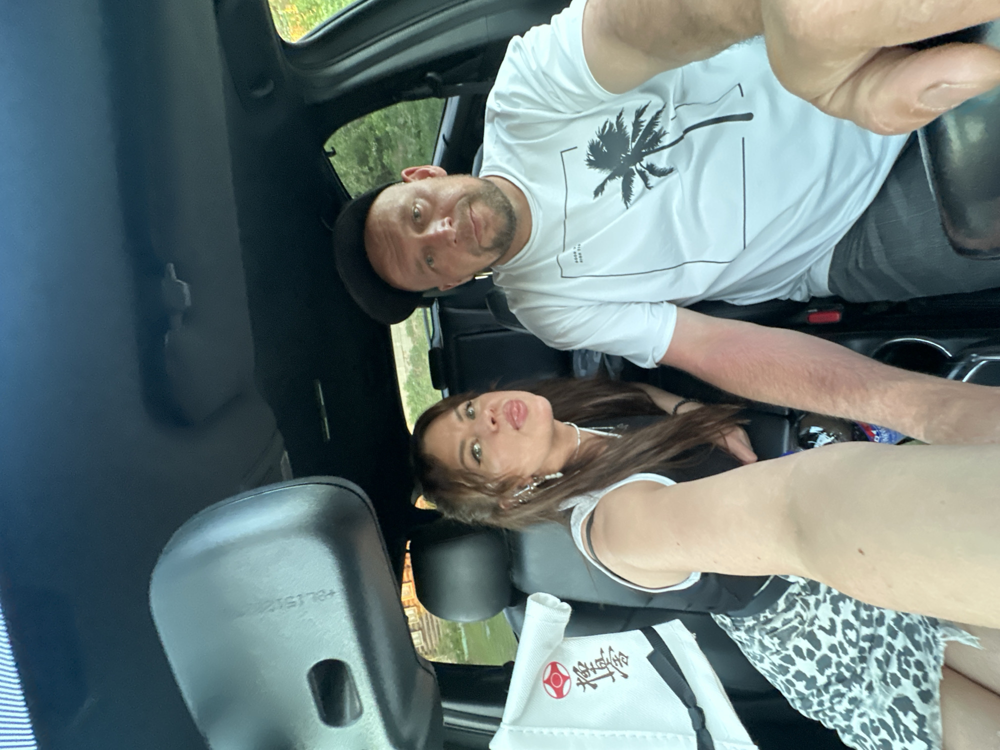 The image shows two people, a man and a woman, inside a car. The man is in the passenger seat, wearing a white t-shirt with a palm tree graphic and a dark cap. The woman is in the driver's seat. She has long brown hair, is wearing a white top and a black and white patterned skirt. A white object with Japanese lettering sits on her lap. The location is not discernible in the image, so the claim that the location is in the city of Chaikovsky, Russia, is not supported.