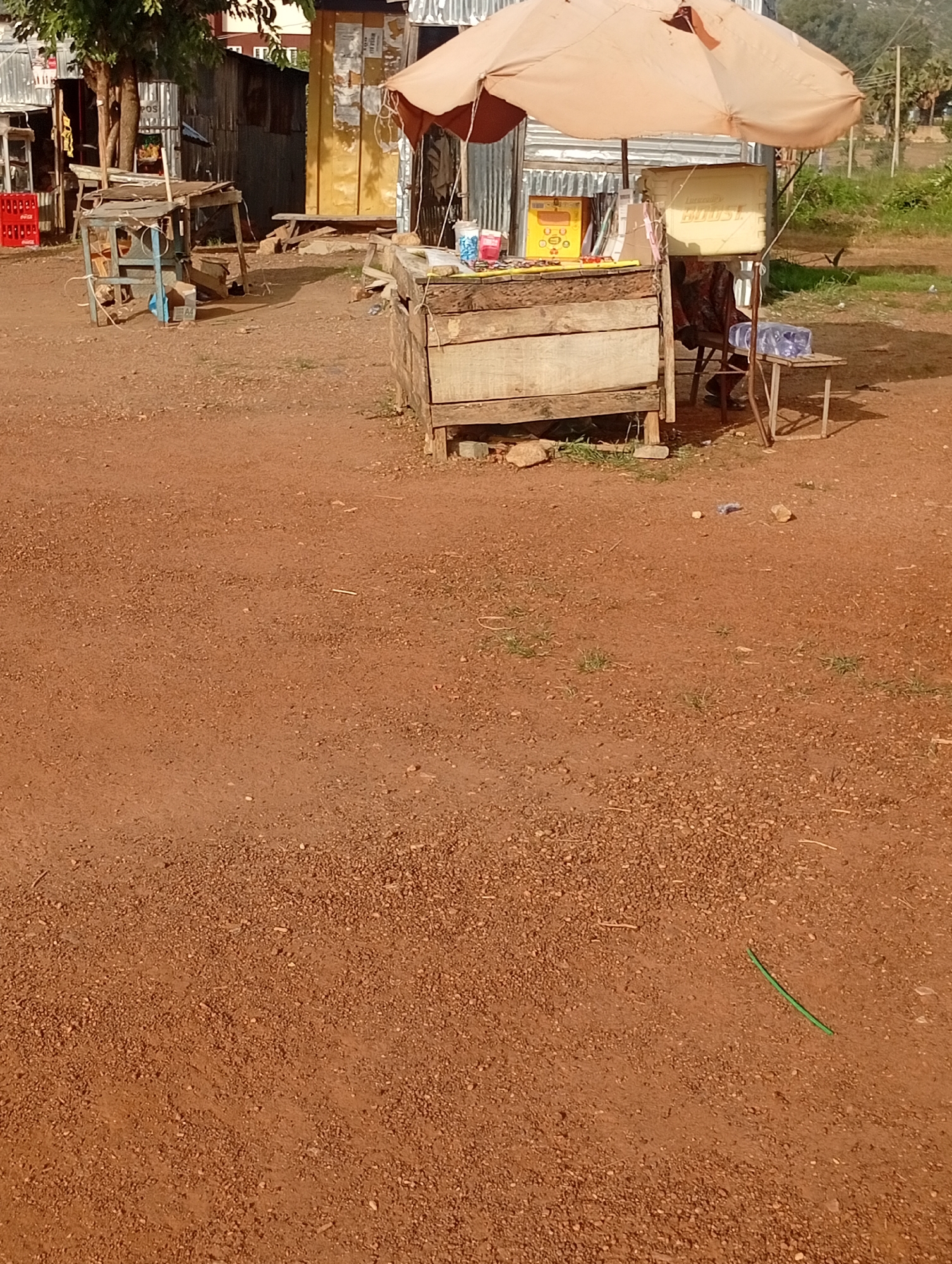 A vendor is selling drinks from a stall shaded by a large umbrella. The stall is made of wood and has several bottles of drinks displayed on it. A cooler box is suspended from the umbrella. A person is sitting on a chair behind the stall. The ground is dirt and gravel. In the background, there are several corrugated iron shacks and some vegetation. The scene appears to be in a market area in Jos, Nigeria, based on the reddish-brown soil common in the region and the typical market setup.